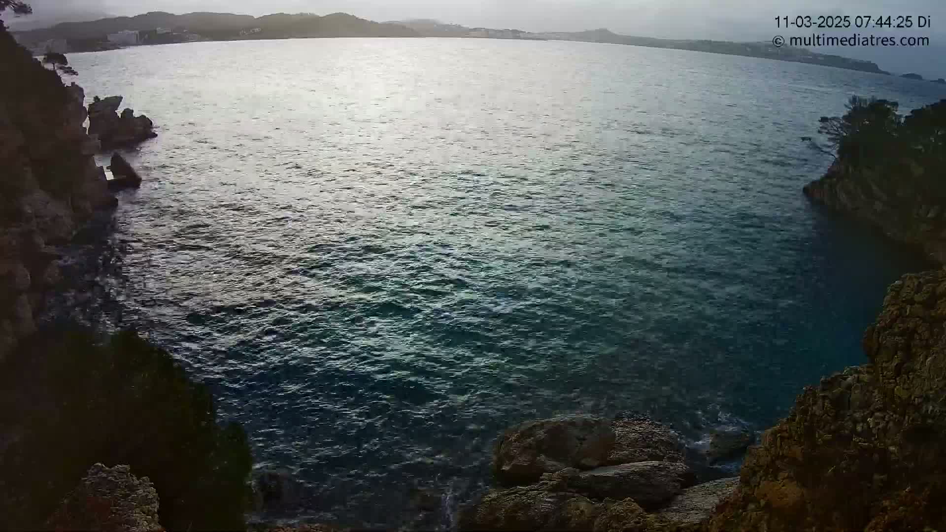 A wide, calm body of water is seen between two rocky shorelines under an overcast sky.