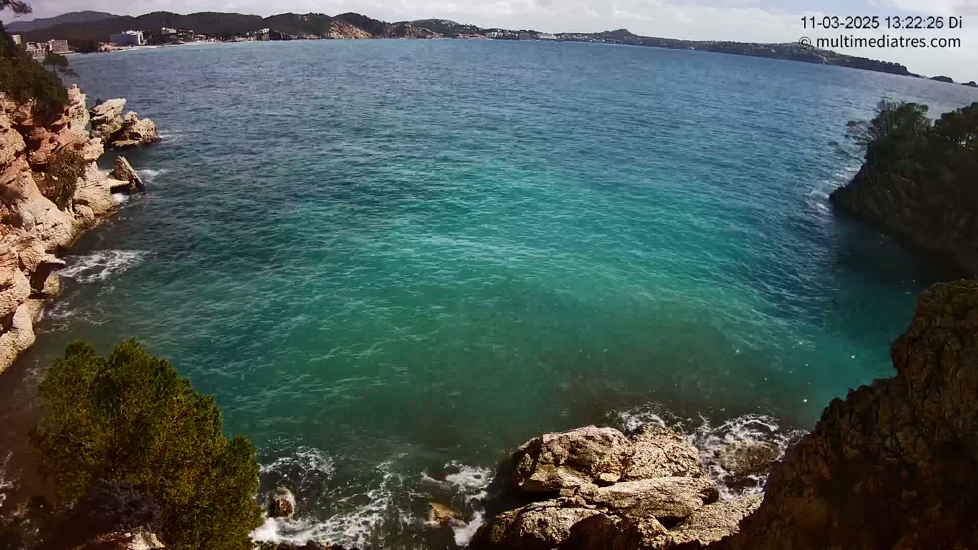 A partly sunny day shows turquoise water lapping against rocky, tan-colored cliffs and a distant coastline.