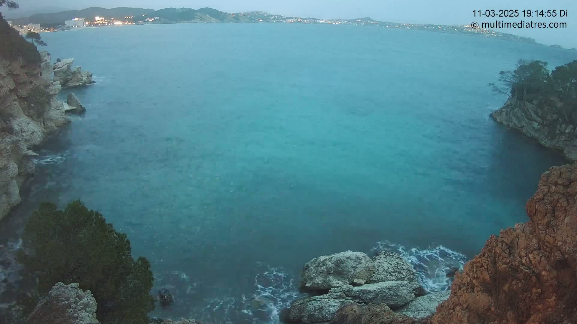 A tranquil teal sea laps against rocky, tree-studded shores under a twilight sky.