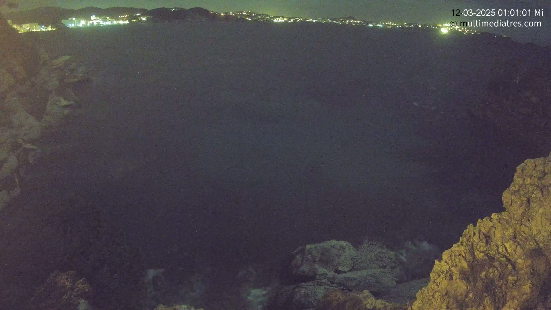 A nighttime view of a dark body of water with a distant illuminated shoreline and rocky outcrops in the foreground.