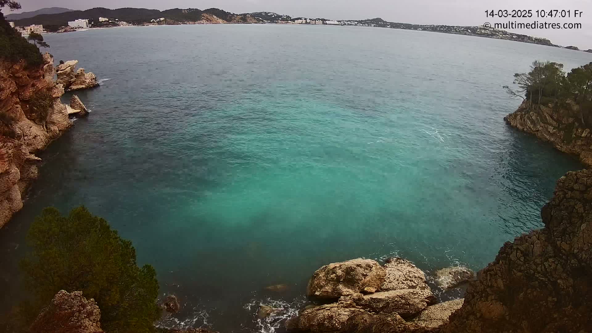 A cove of teal water is nestled between rocky cliffs on an overcast day, with a distant coastline visible in the background.