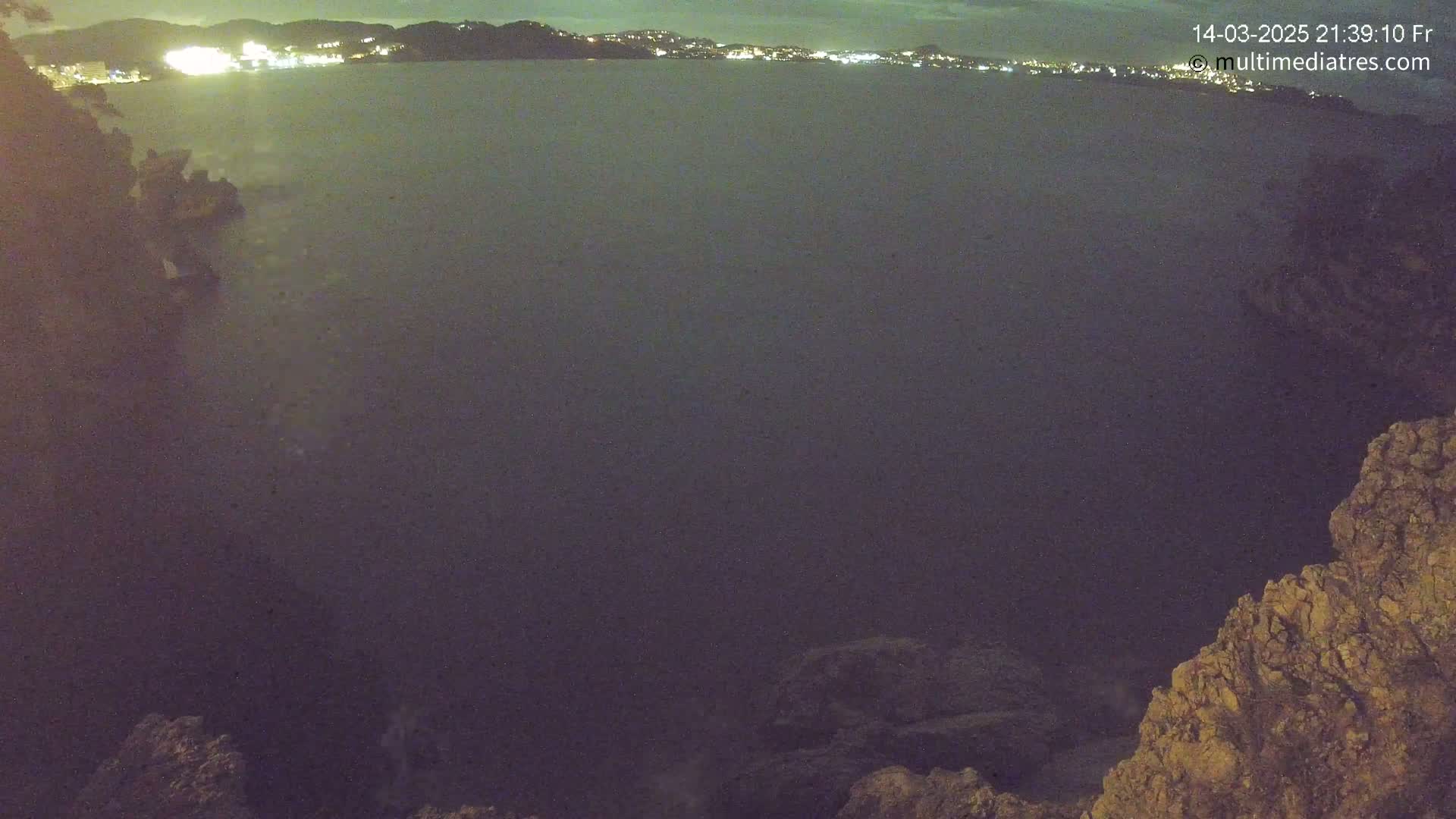 A nighttime view from a rocky outcrop shows a dark body of water with a distant, faintly lit coastline.