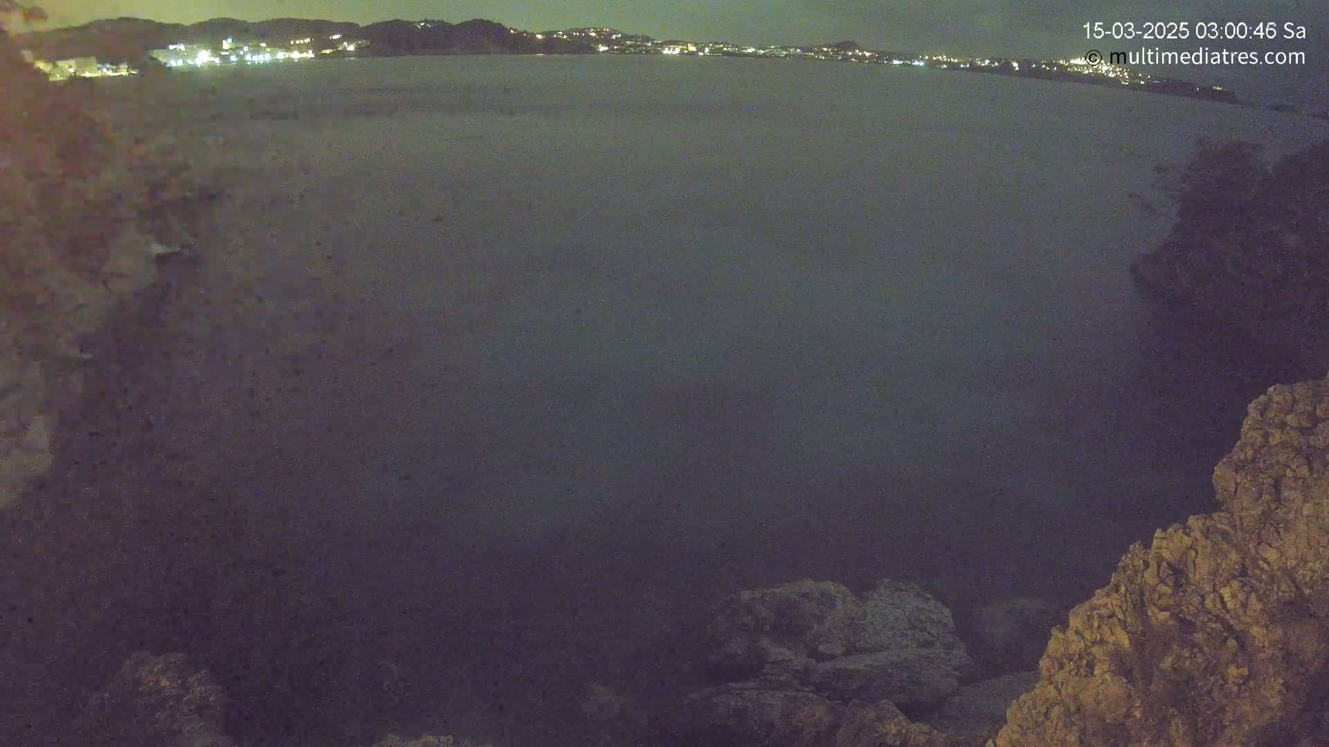 A dark, nighttime view of a calm body of water with a distant city skyline illuminated, seen from a rocky shoreline.