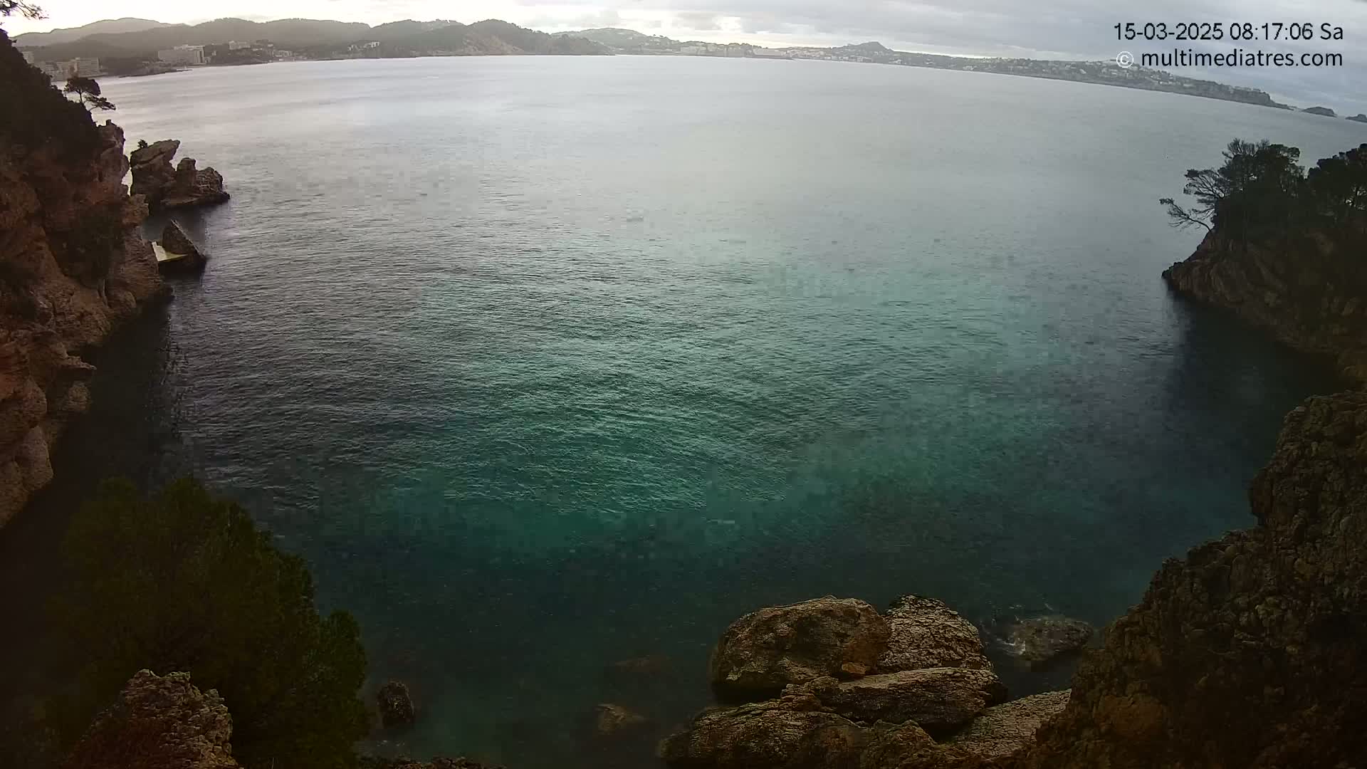 A cove of dark teal water is nestled between rocky cliffs under a cloudy sky.
