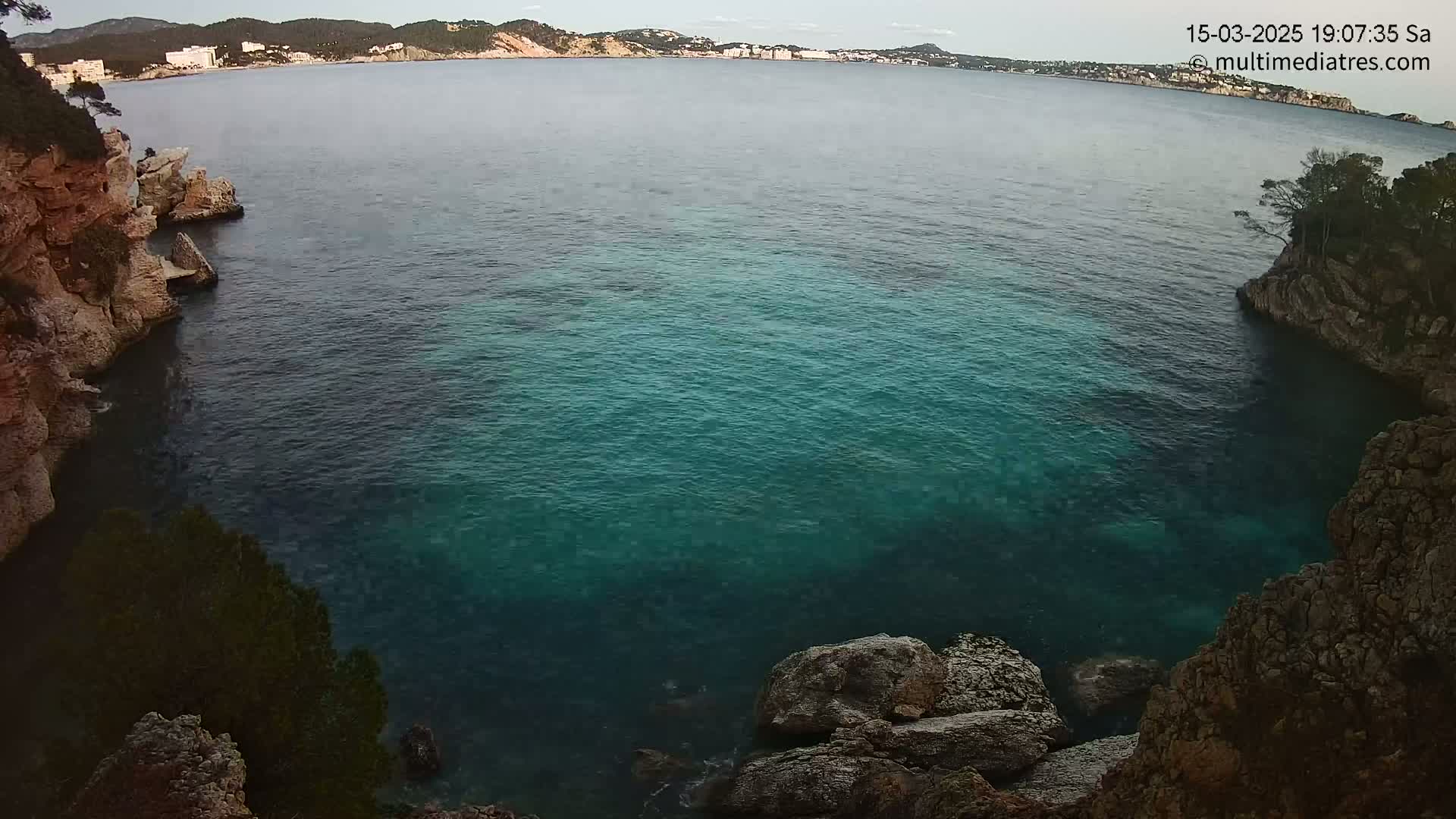 A cove of teal water is nestled between rocky cliffs under a mostly clear sky.