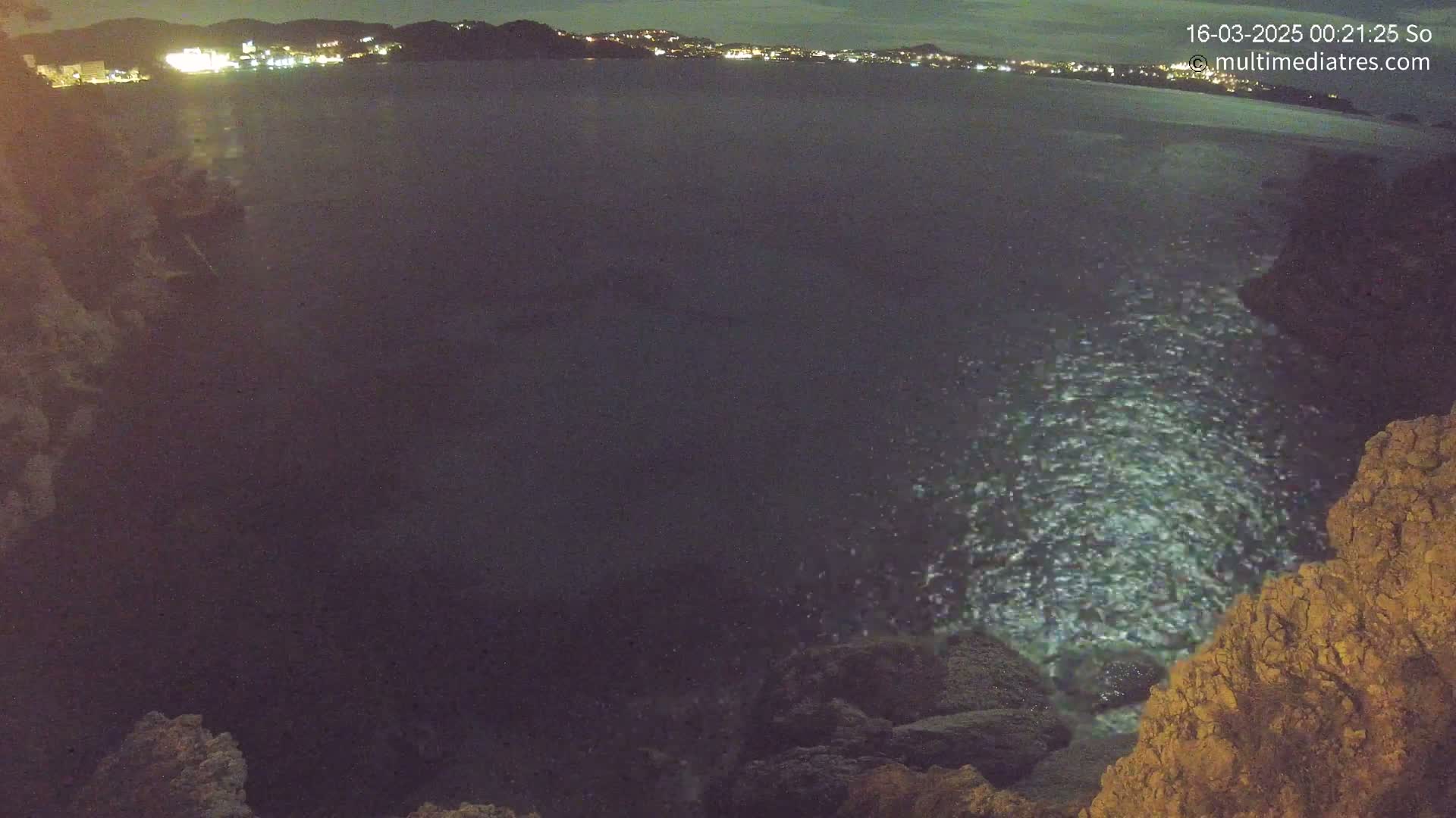 A dark, nighttime view of a rocky coastline with a calm sea, illuminated by distant lights on shore.