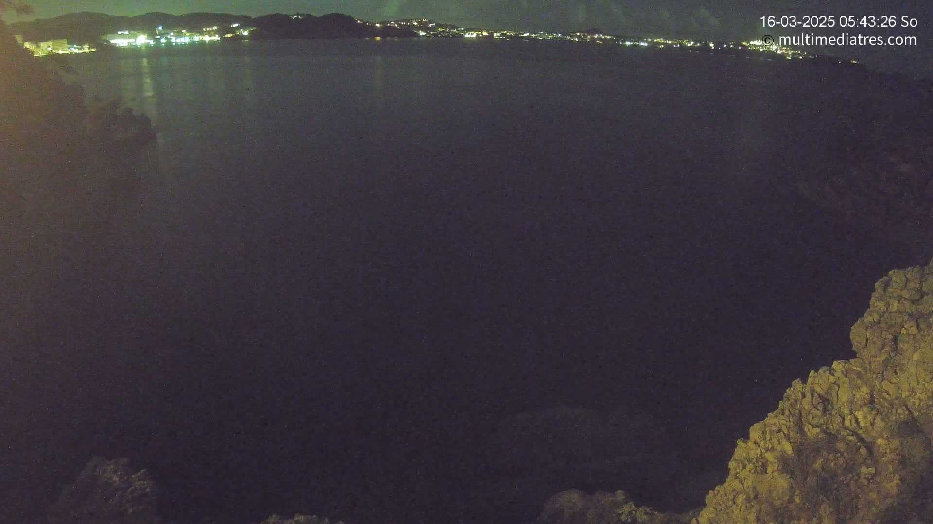 A dark body of water at night is seen from a high vantage point, with a distant, dimly lit town visible across the water and a rocky outcrop in the foreground.