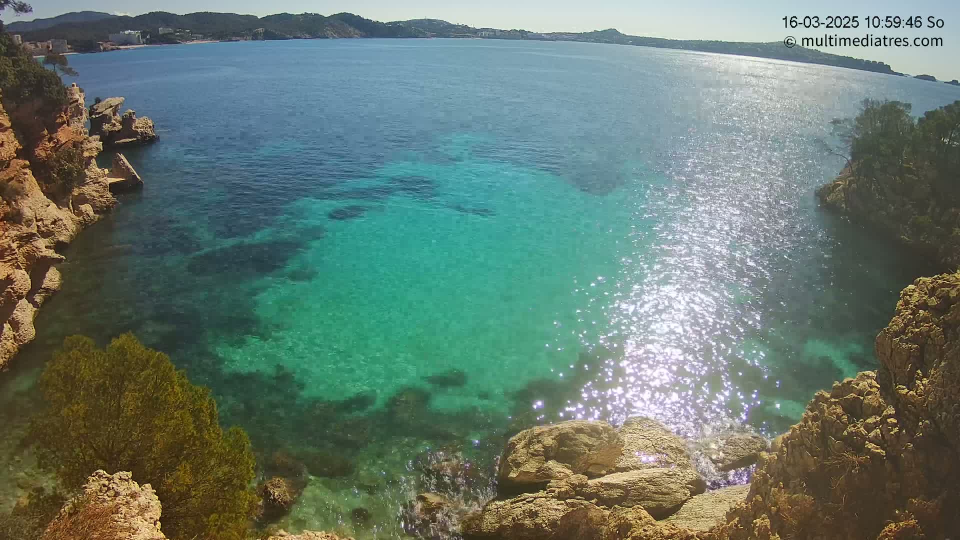 Sunlight glistens on clear, turquoise water surrounding rocky, tan cliffs on a sunny day.