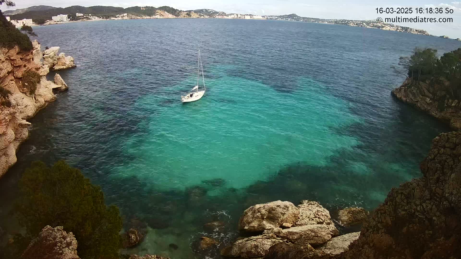 A sailboat sits in a calm, teal cove surrounded by rocky, light brown cliffs under a partly cloudy sky.