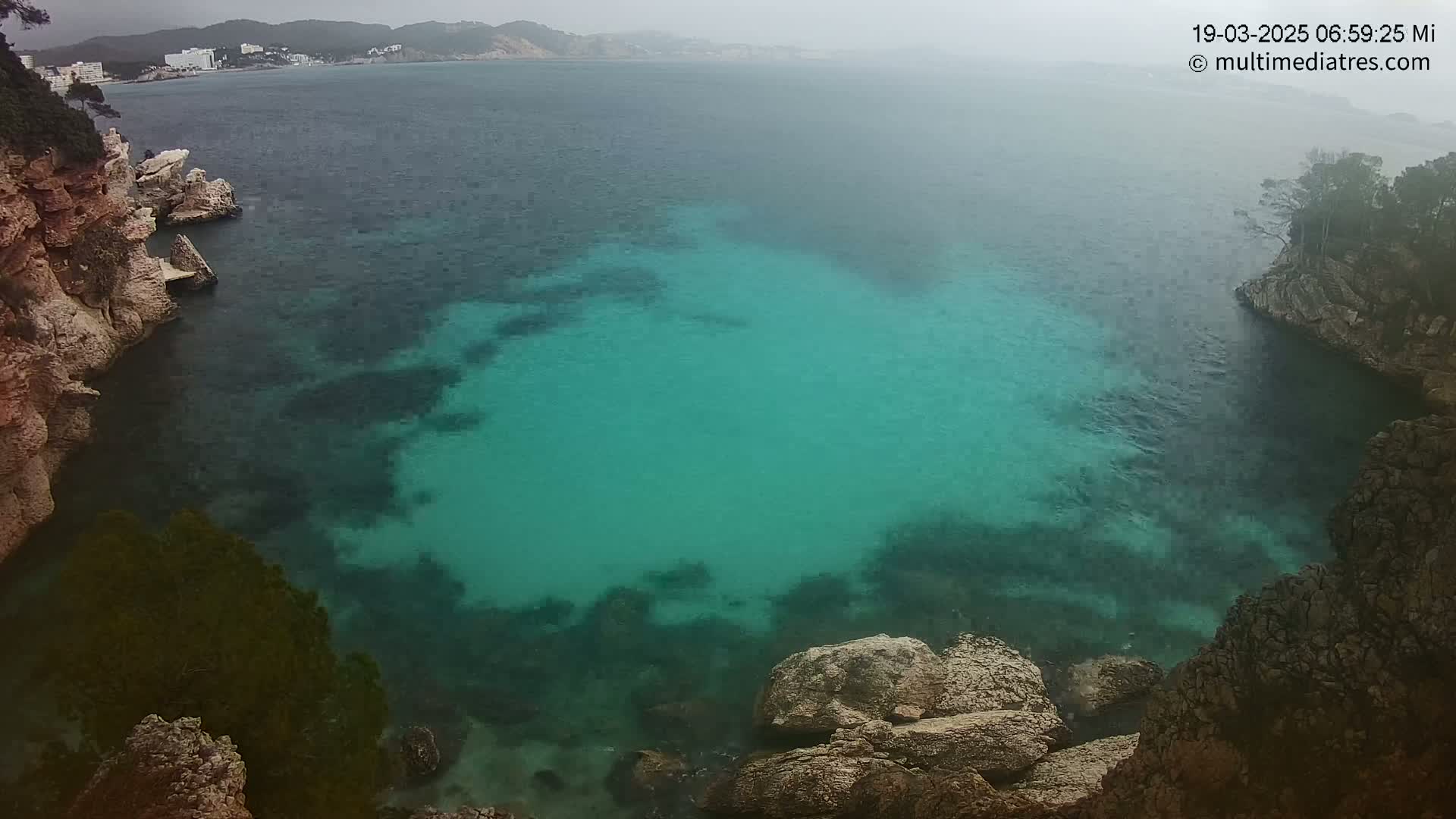 A cove of clear, teal water is nestled between rocky cliffs on an overcast day, with a distant coastline visible in the background.