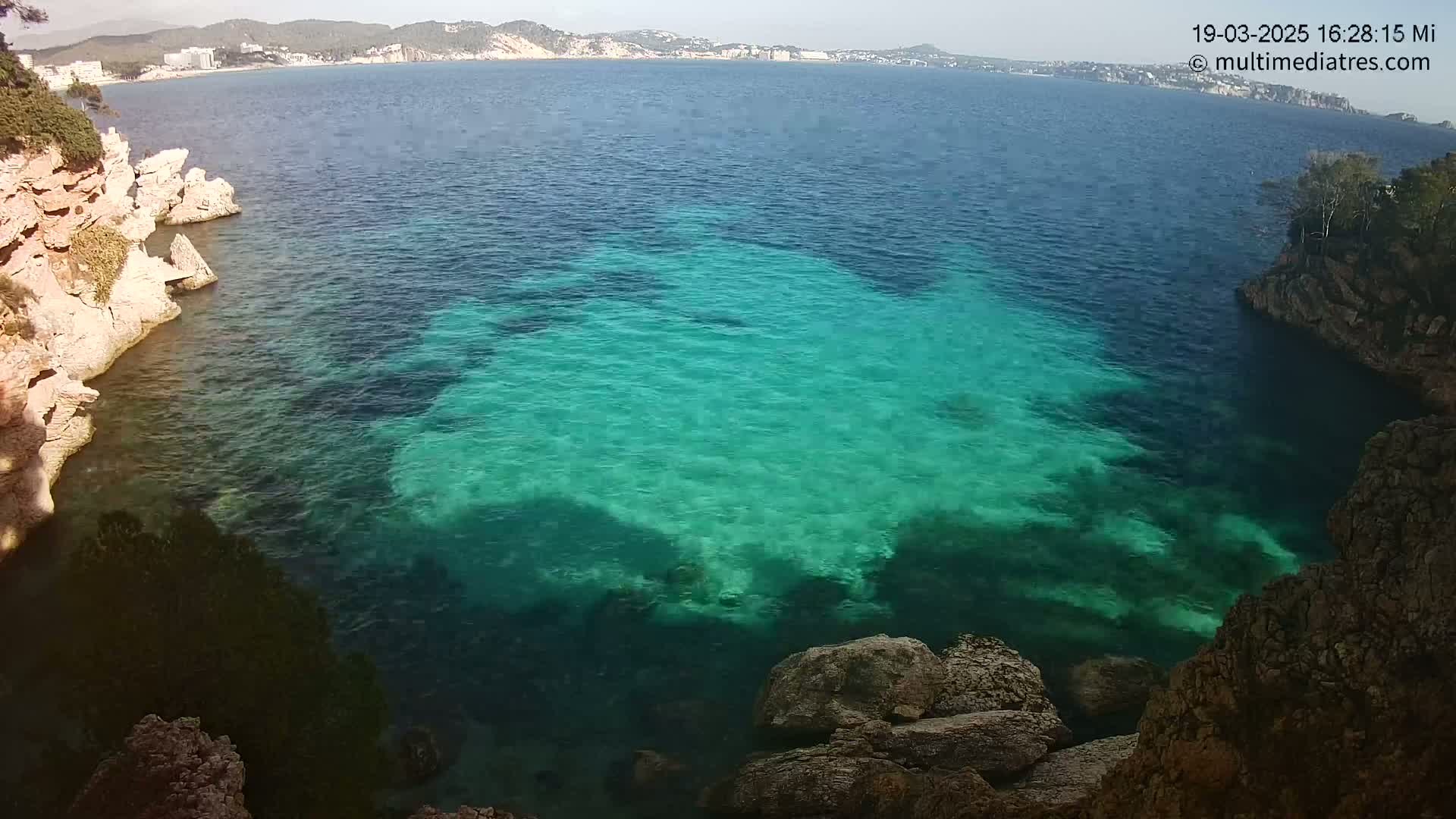 Clear, teal water fills a cove surrounded by light-brown, rocky cliffs, with a distant coastline visible under a sunny sky.