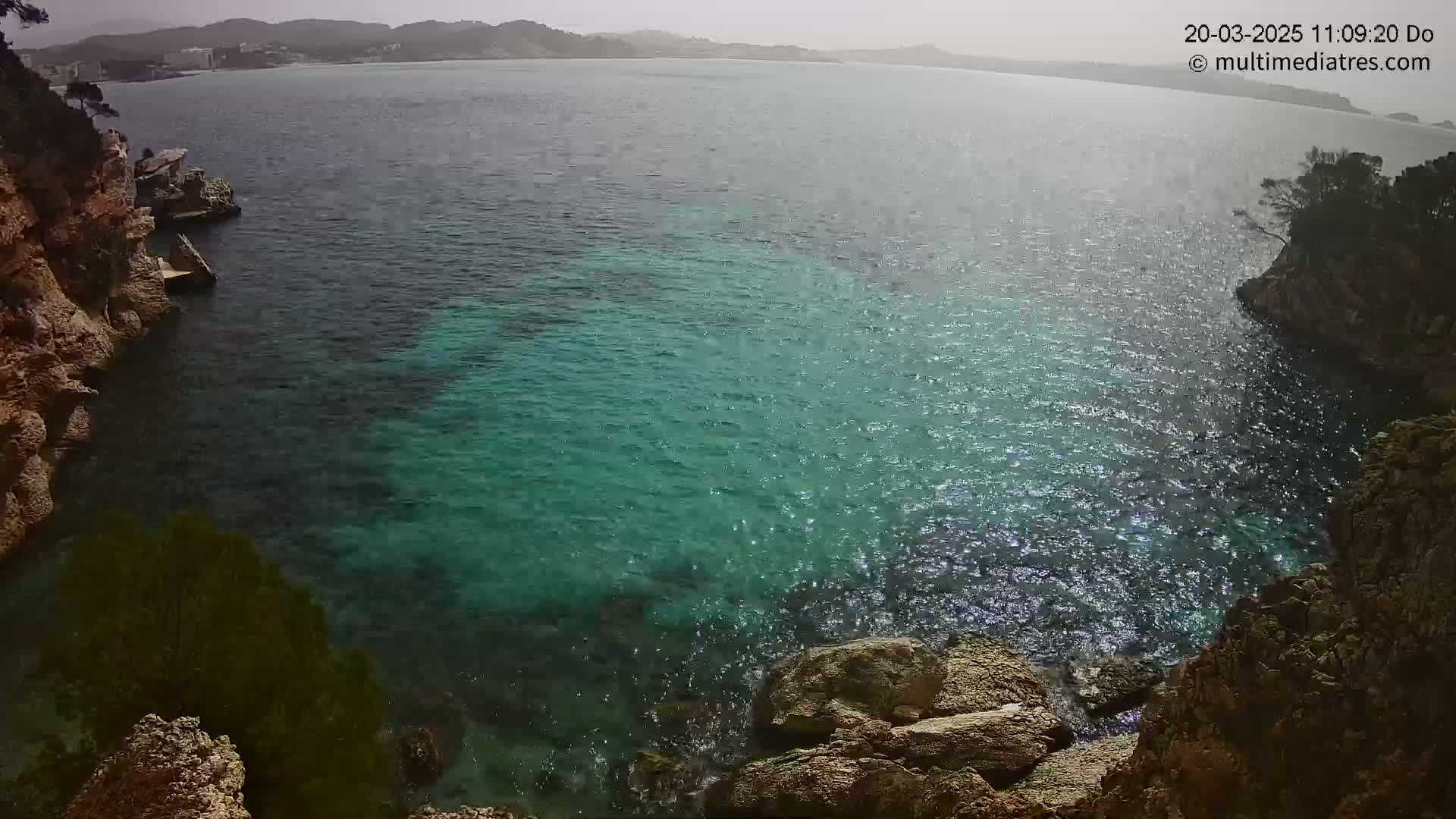 A rocky cove with teal water stretches to a hazy coastline under an overcast sky.