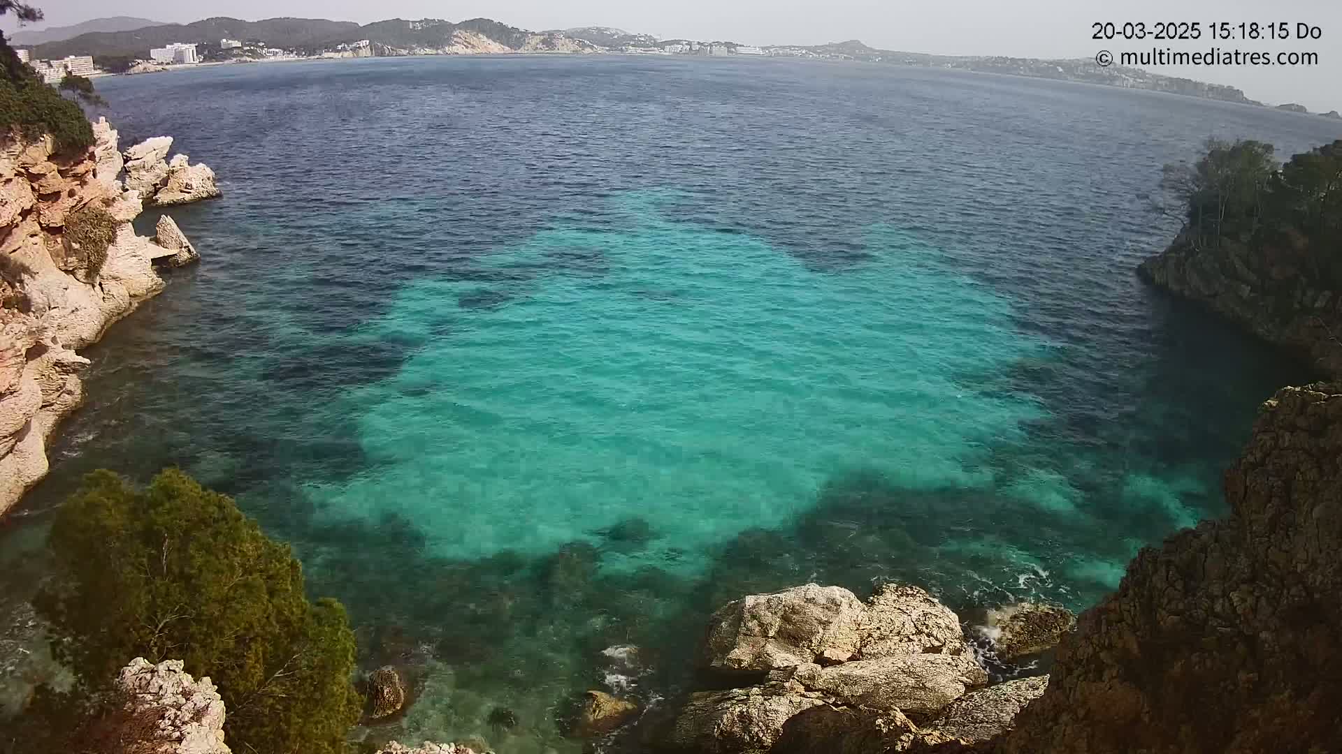 A clear, bright cove of teal water is nestled between rocky, light-brown cliffs on a sunny day.