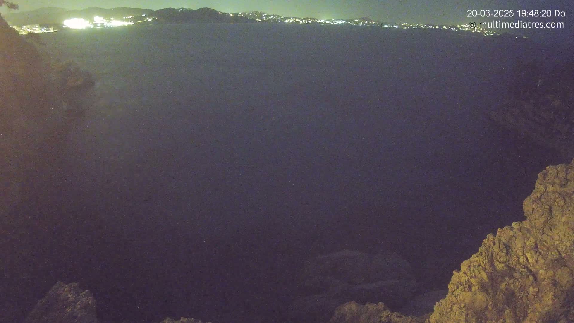 A dark body of water at night is viewed from a rocky outcrop, with a distant city's lights visible on the horizon.