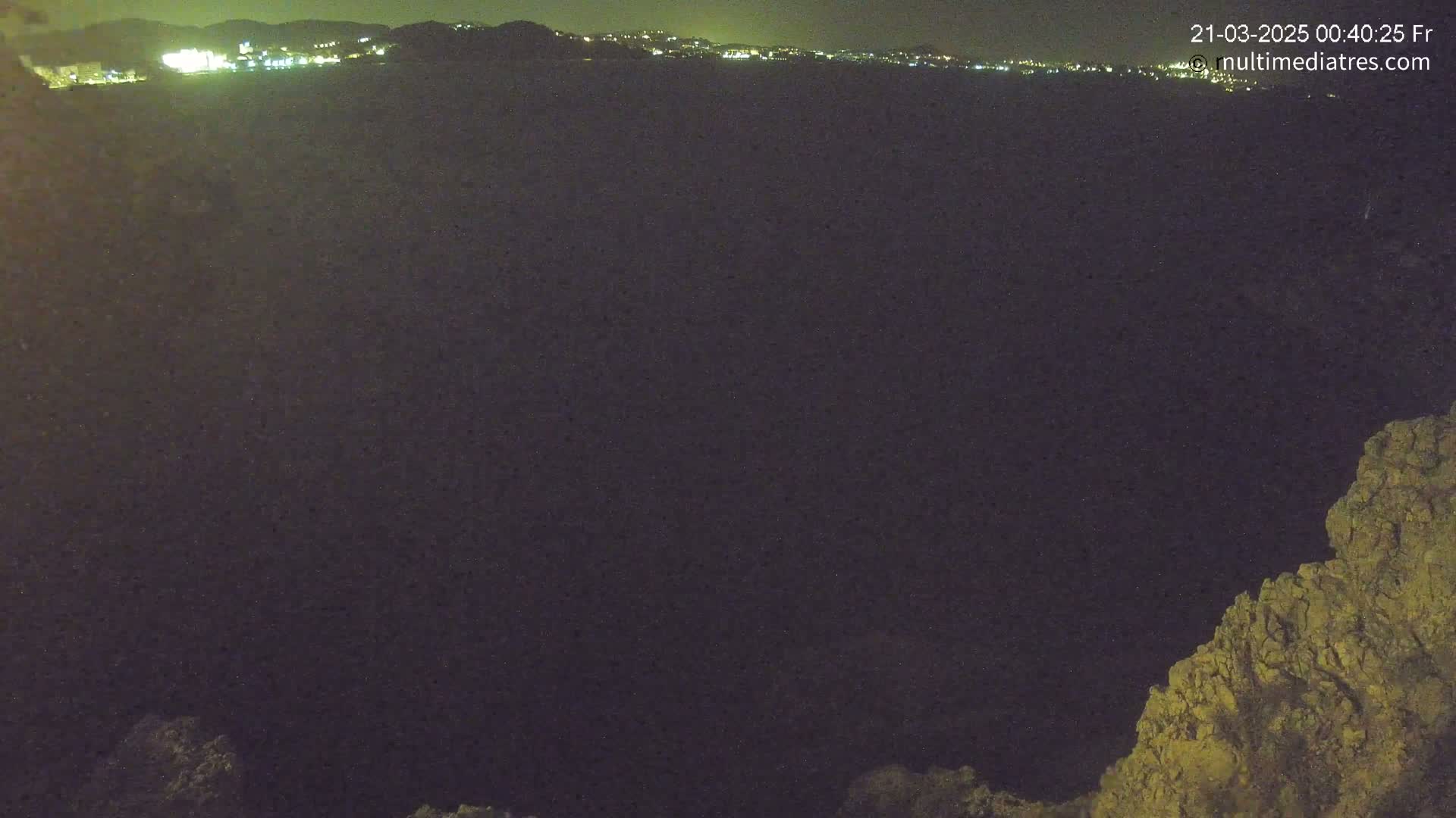 A nighttime view of a dark, possibly mountainous area, with a distant city skyline faintly illuminated in the background.