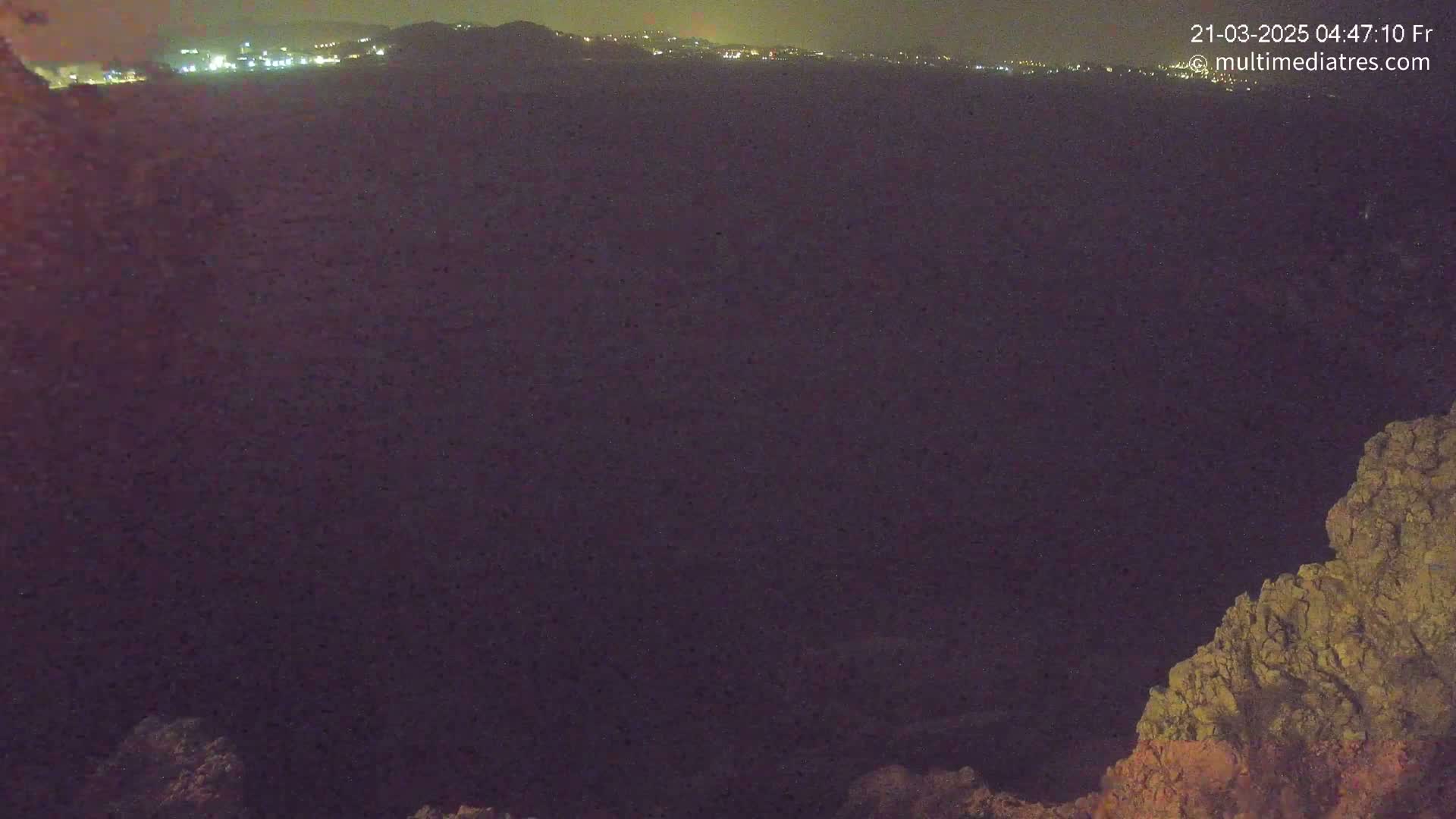 A nighttime long shot shows a dark, possibly mountainous landscape with a distant line of lights and a rocky foreground under a clear, dark sky.