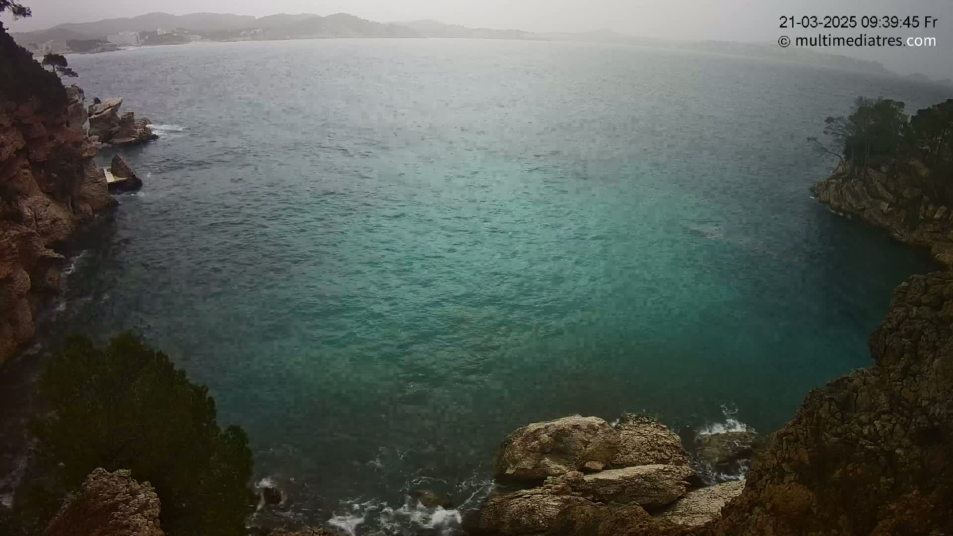 A view from above shows a teal-colored cove surrounded by rocky cliffs and trees under an overcast sky.