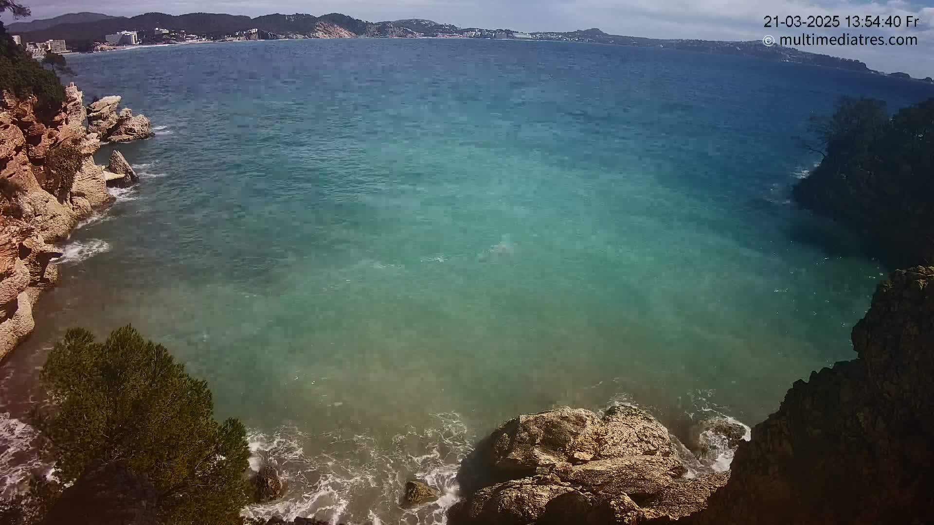 A cove of teal-colored water, bordered by light brown rocky cliffs and small waves lapping at the shore, with a distant view of a town under a partly cloudy sky.