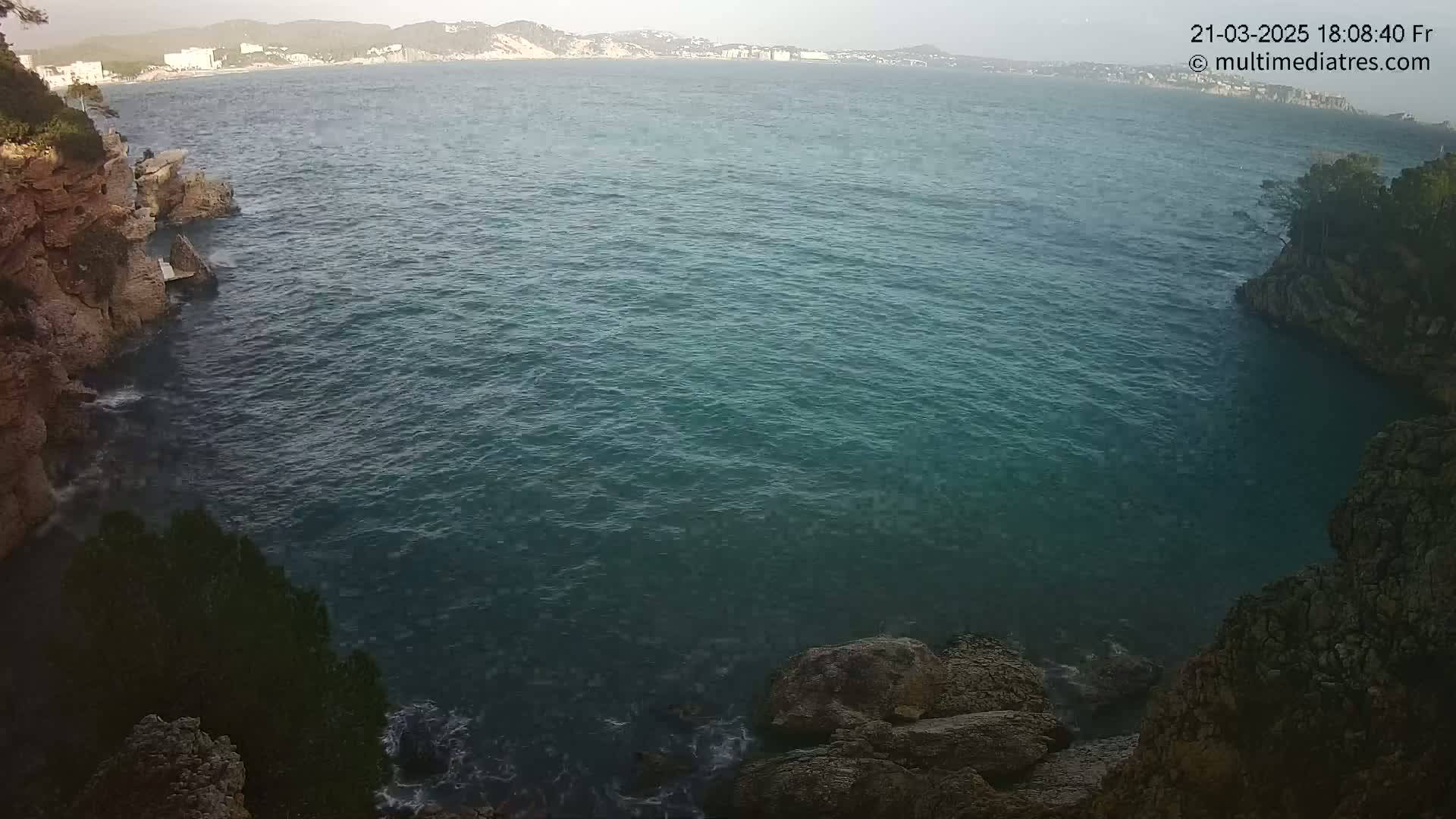 A calm, dark-blue sea is seen between rocky cliffs, with a distant shoreline visible in the background under a clear sky.