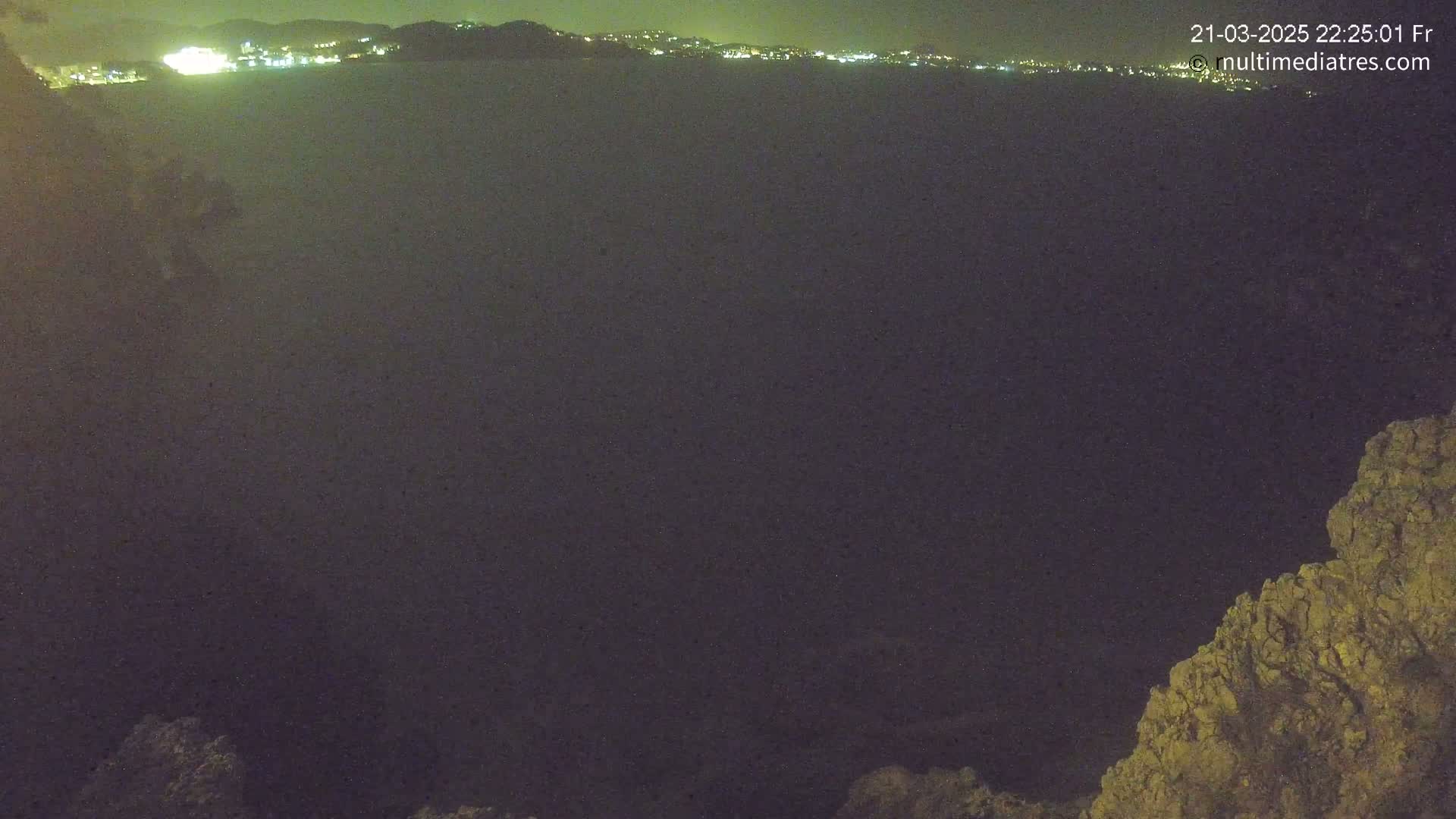 A nighttime, dark view of a body of water with a distant, illuminated city skyline in the background and rocky terrain in the foreground.