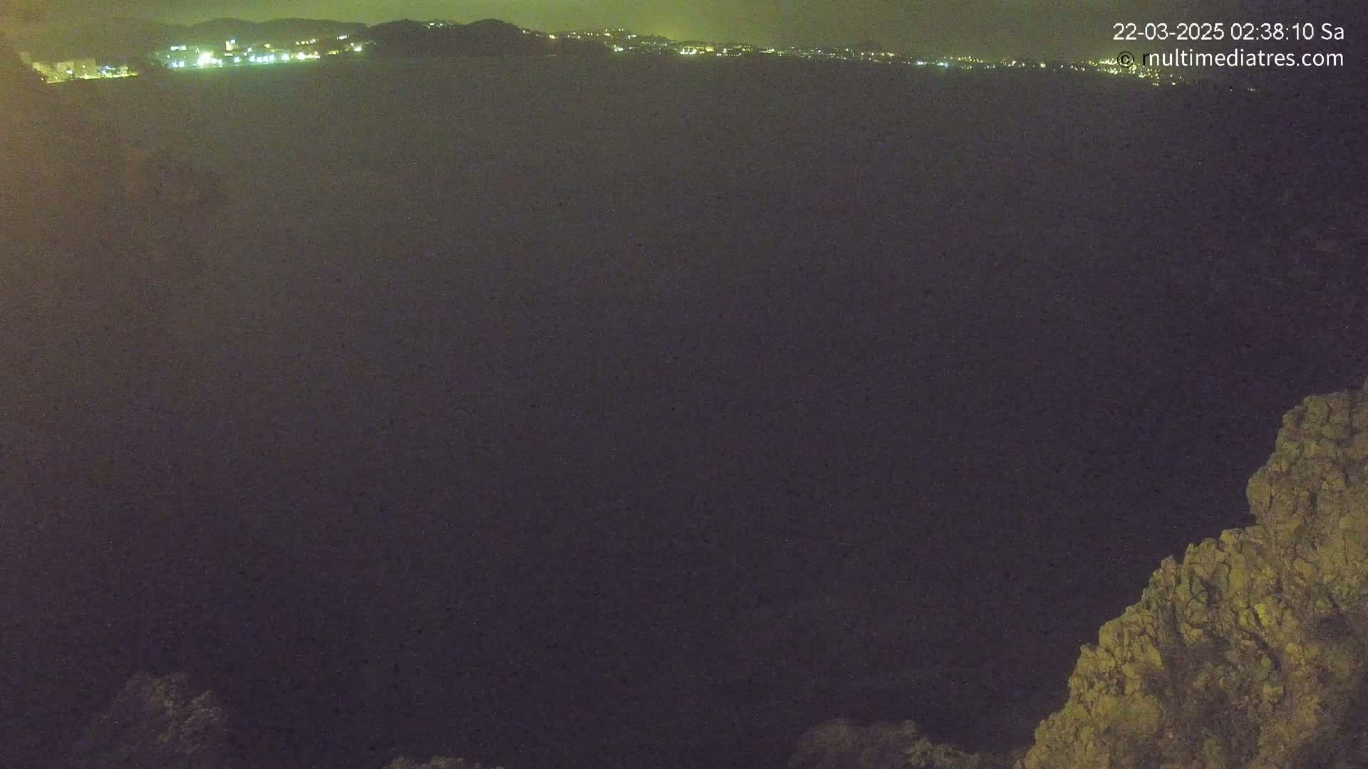 A nighttime, dark view shows a body of water with a distant line of city lights, and a rocky outcrop in the lower right corner.