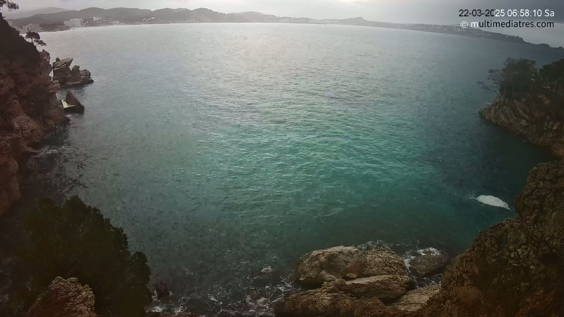 A tranquil teal sea is nestled between rocky cliffs on an overcast day, with a distant shoreline visible in the background.