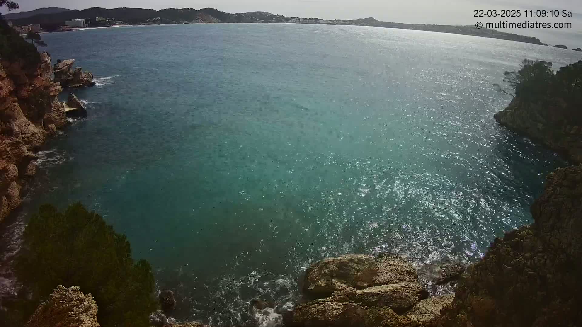 A partly sunny day shows a rocky cove with teal water lapping against the shore, extending to a calm bay with a distant shoreline.