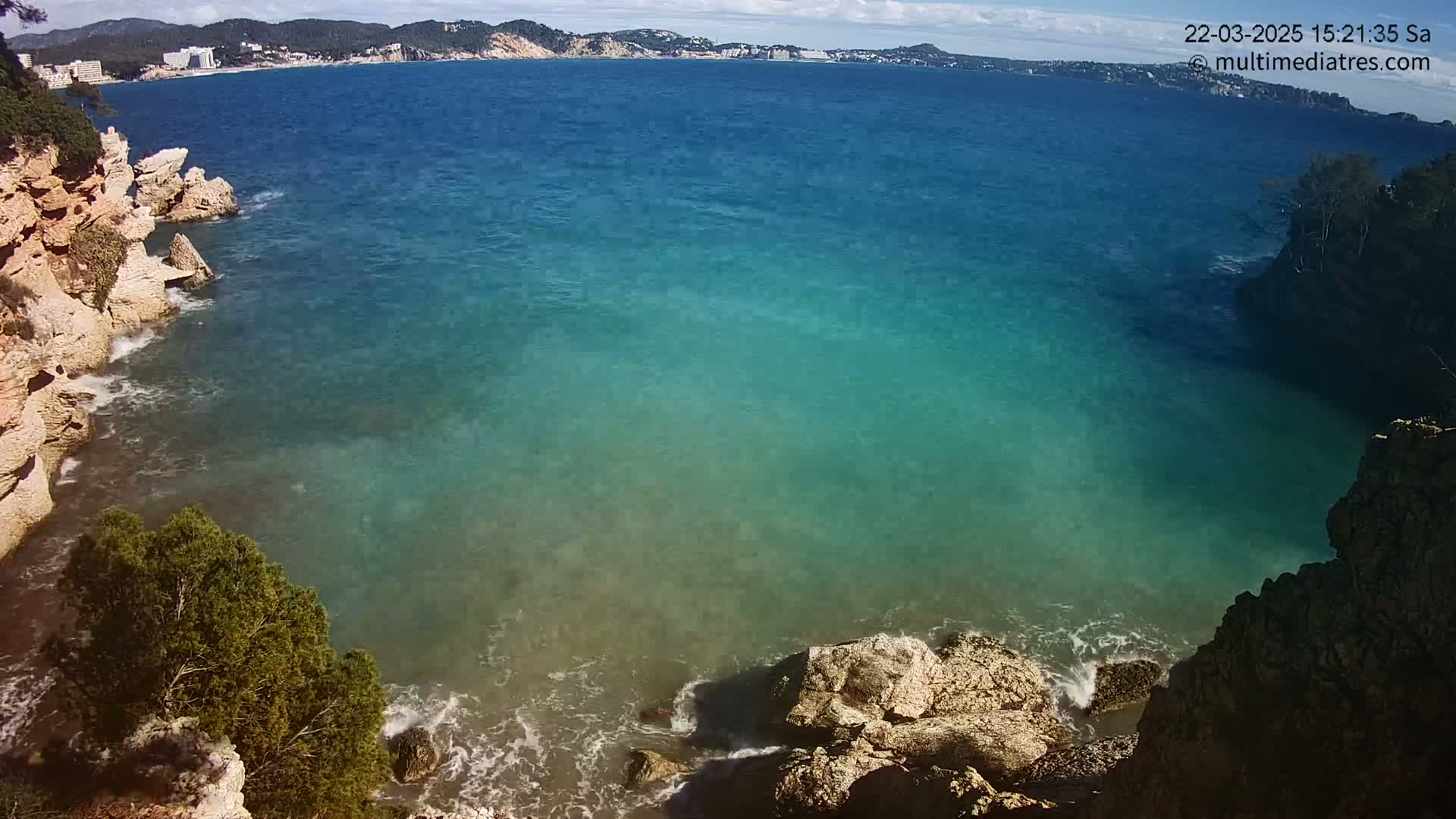 A cove of teal water is surrounded by light-brown rocky cliffs and small trees, under a partly sunny sky.