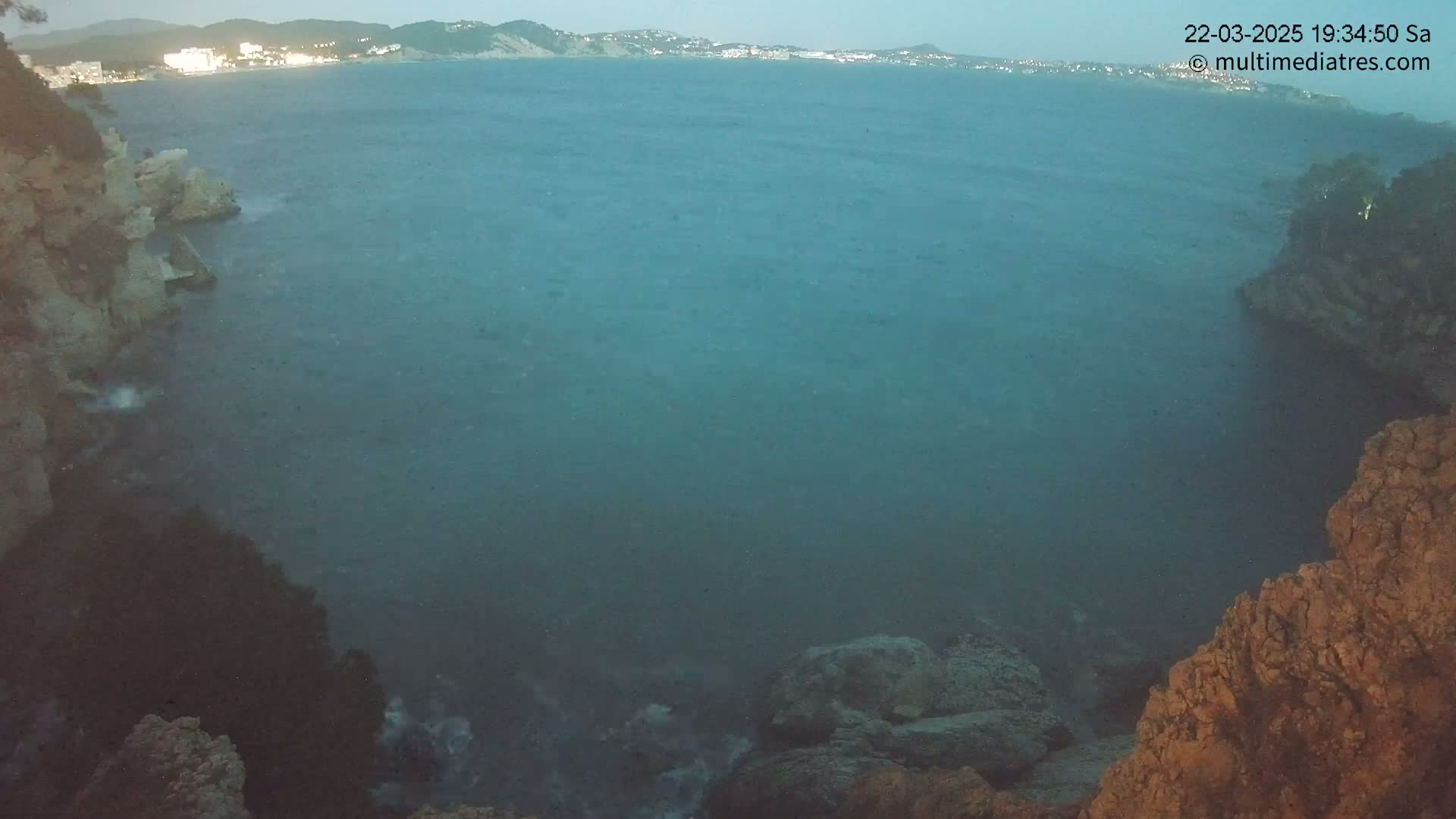 A dark-blue body of water, slightly choppy, is seen from a high vantage point between rocky shores, with a distant town visible on the far shore under a twilight sky.