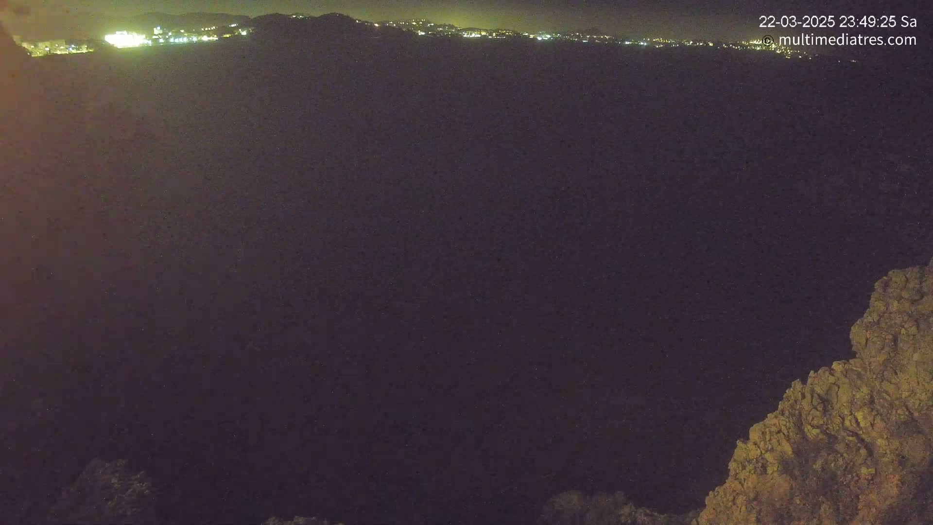 A nighttime view shows a dark expanse, possibly a valley or plain, with a distant line of city lights and a rocky outcrop in the foreground under a clear, dark sky.