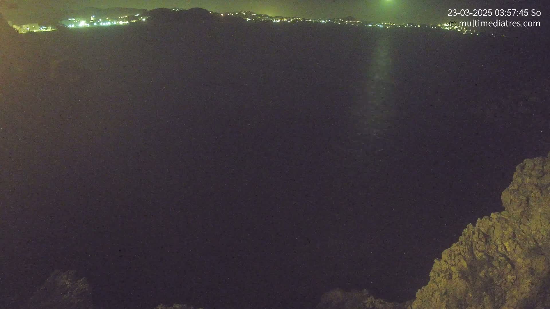 A nighttime view shows a dark expanse, possibly a valley or crater, with a distant line of city lights under a clear, dark sky, and a rocky outcrop in the foreground at the bottom right.