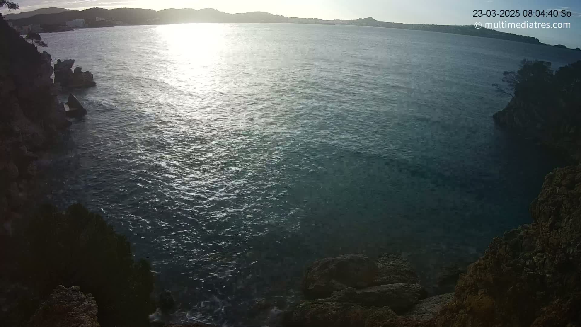 A sunlit body of water is seen from a rocky, high vantage point, with calm waves lapping against the shore.