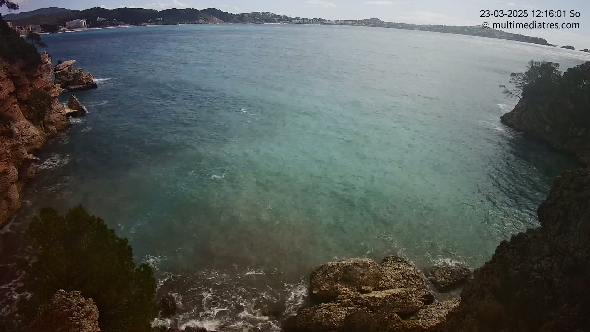A rocky cove with clear teal water meets a calm sea under a partly cloudy sky.