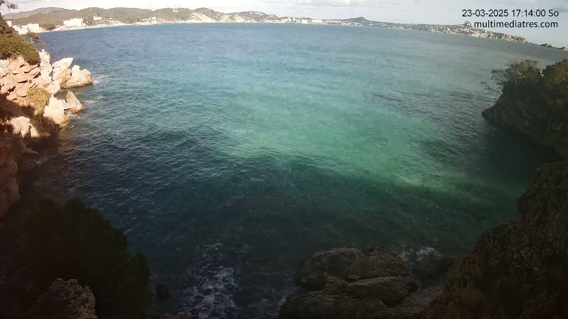A cove with calm, teal-green water is surrounded by rocky cliffs and overlooks a distant coastline under a partly cloudy sky.