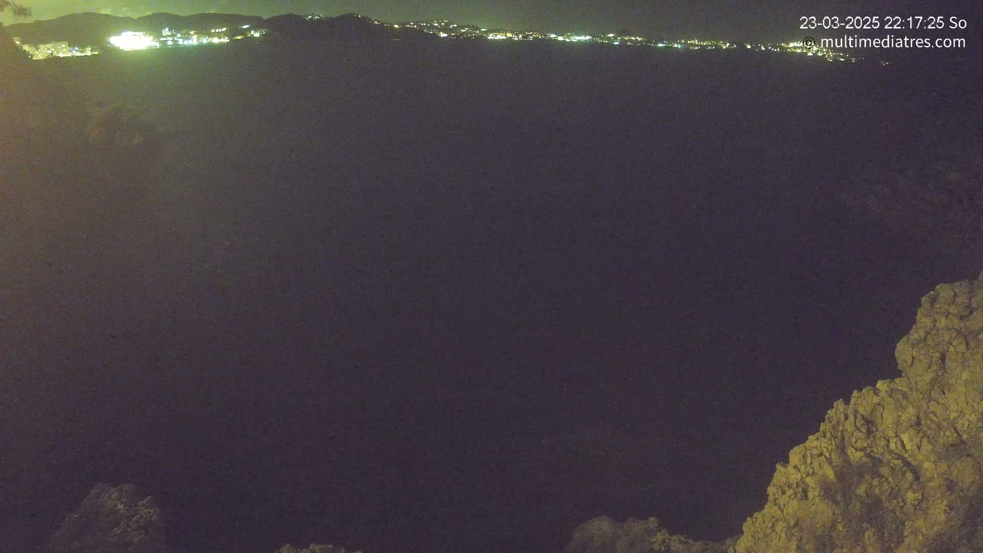 A nighttime long shot shows a distant city skyline illuminated against a dark, clear sky, with rocky terrain visible in the bottom right corner.