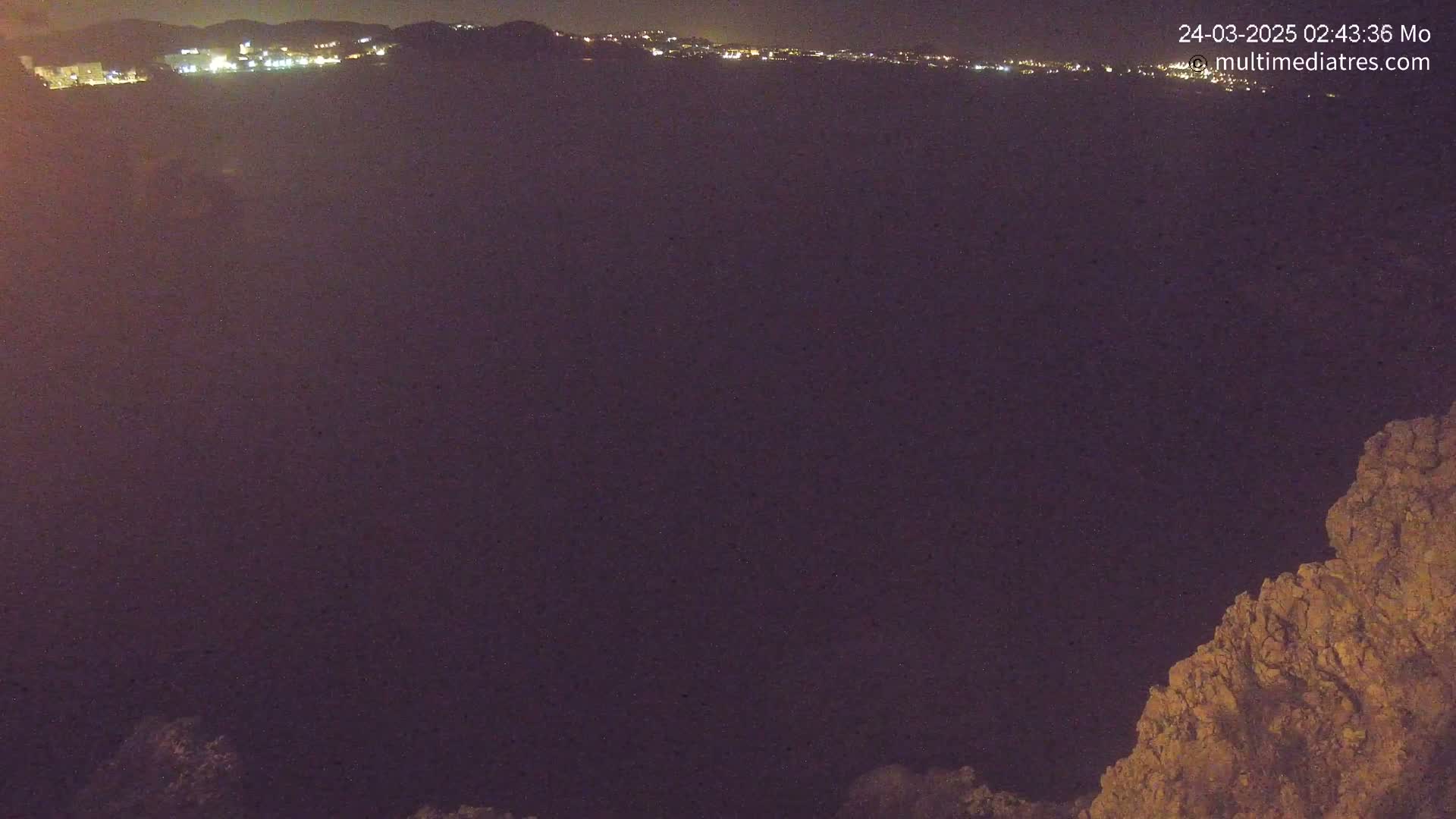 A nighttime, long shot shows a dark expanse of land with a distant line of lights from a town or city in the background, and a rocky outcrop in the bottom right corner.