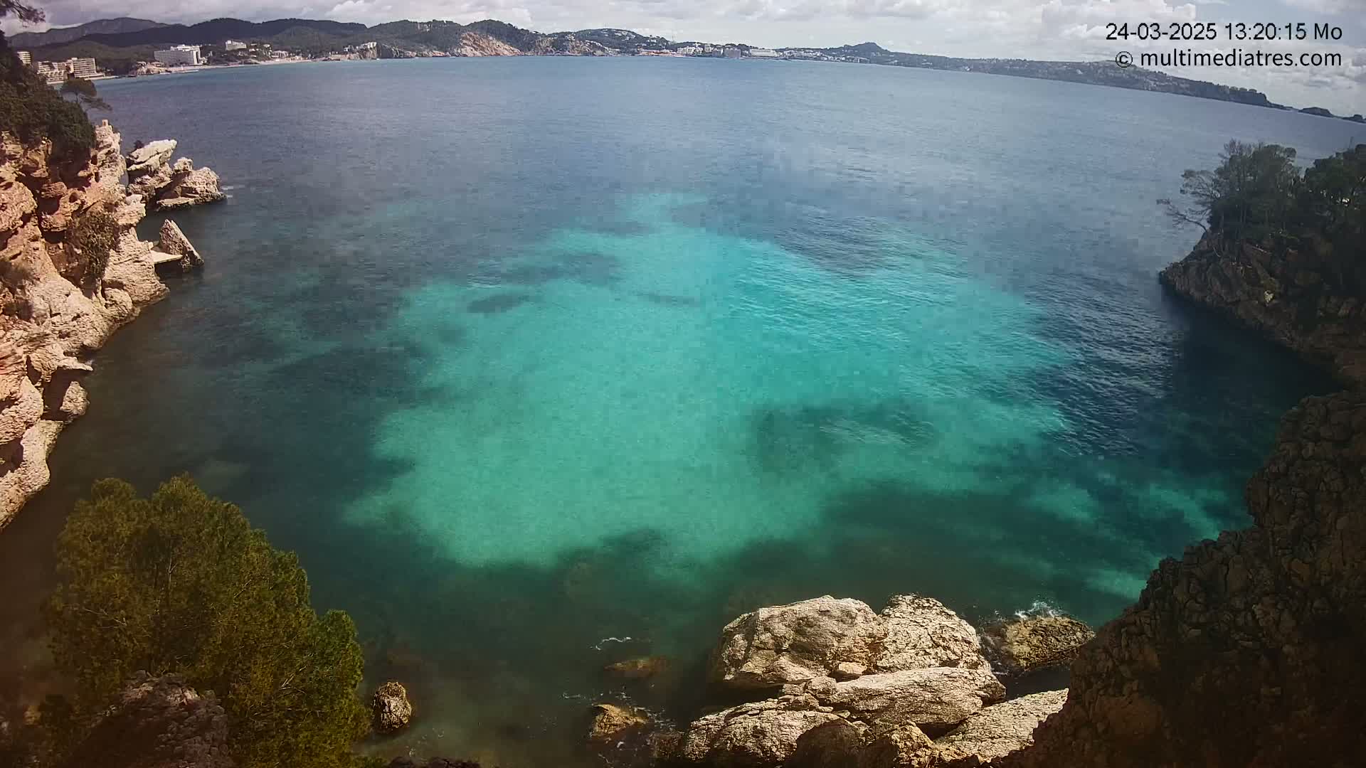A partly cloudy day reveals a cove of teal water nestled between rocky, tree-lined shores, with a distant coastline visible in the background.