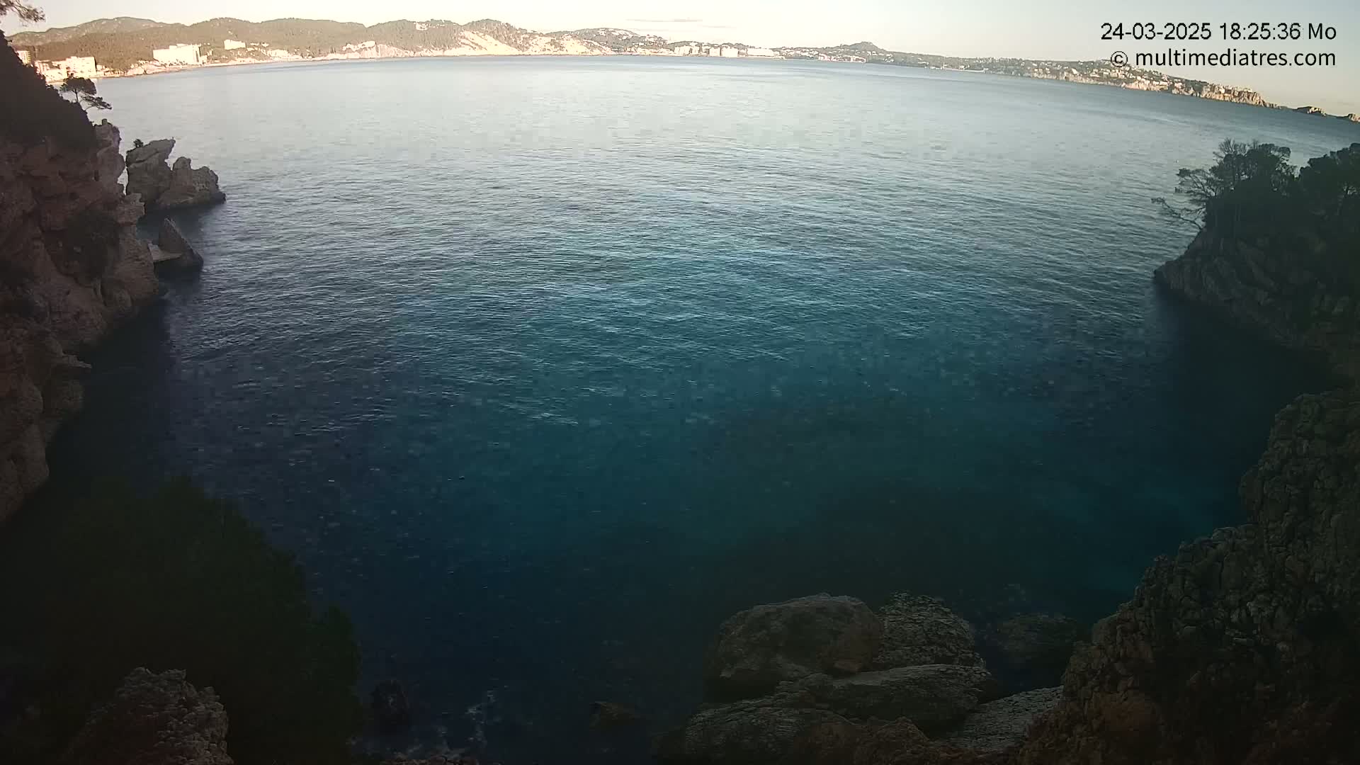 A calm, dark-blue sea is nestled between rocky, cliff-like shores under a clear evening sky.
