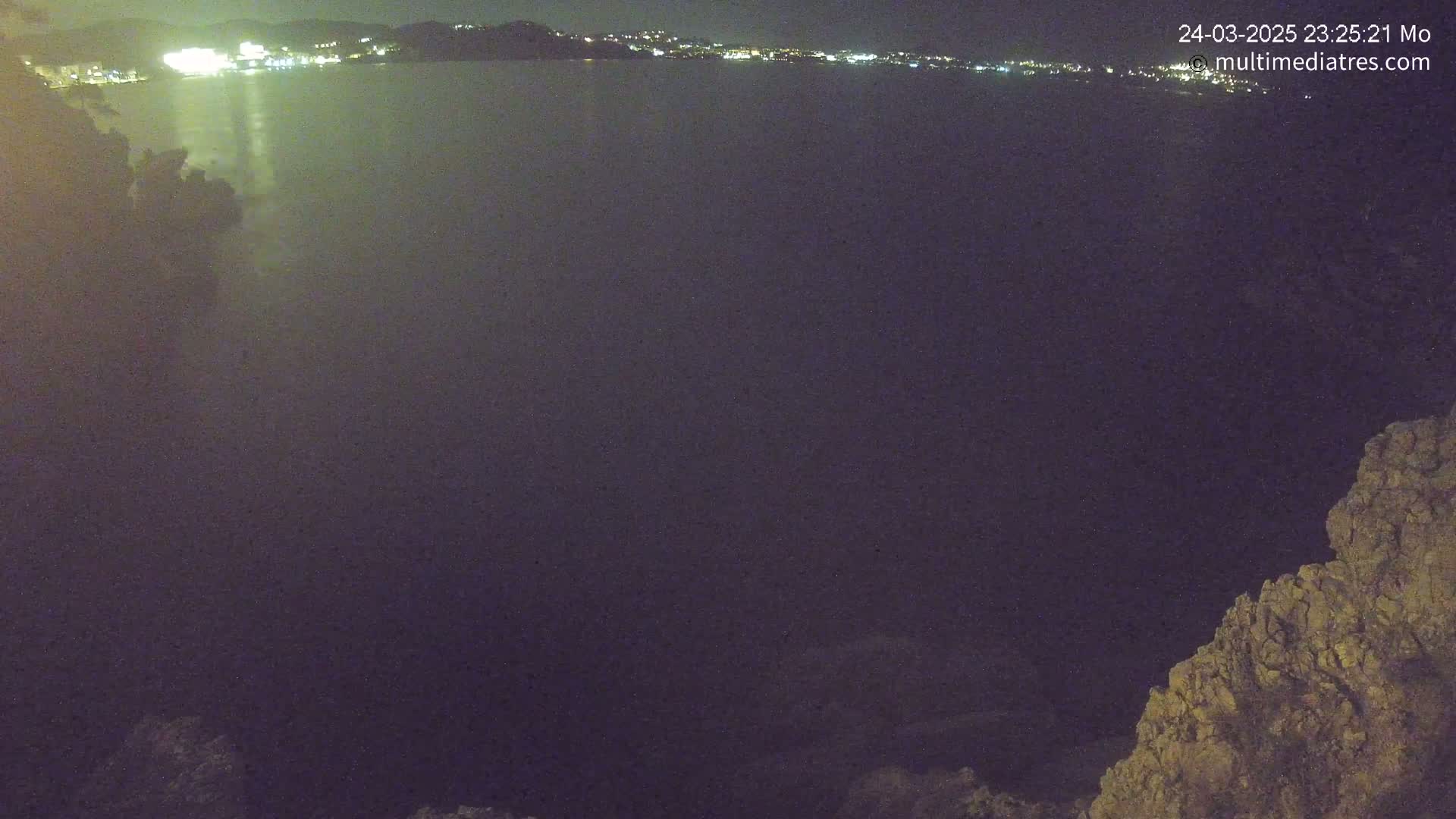 A nighttime view shows a dark body of water reflecting the lights of a city on the horizon, with a rocky outcrop in the foreground under a clear night sky.