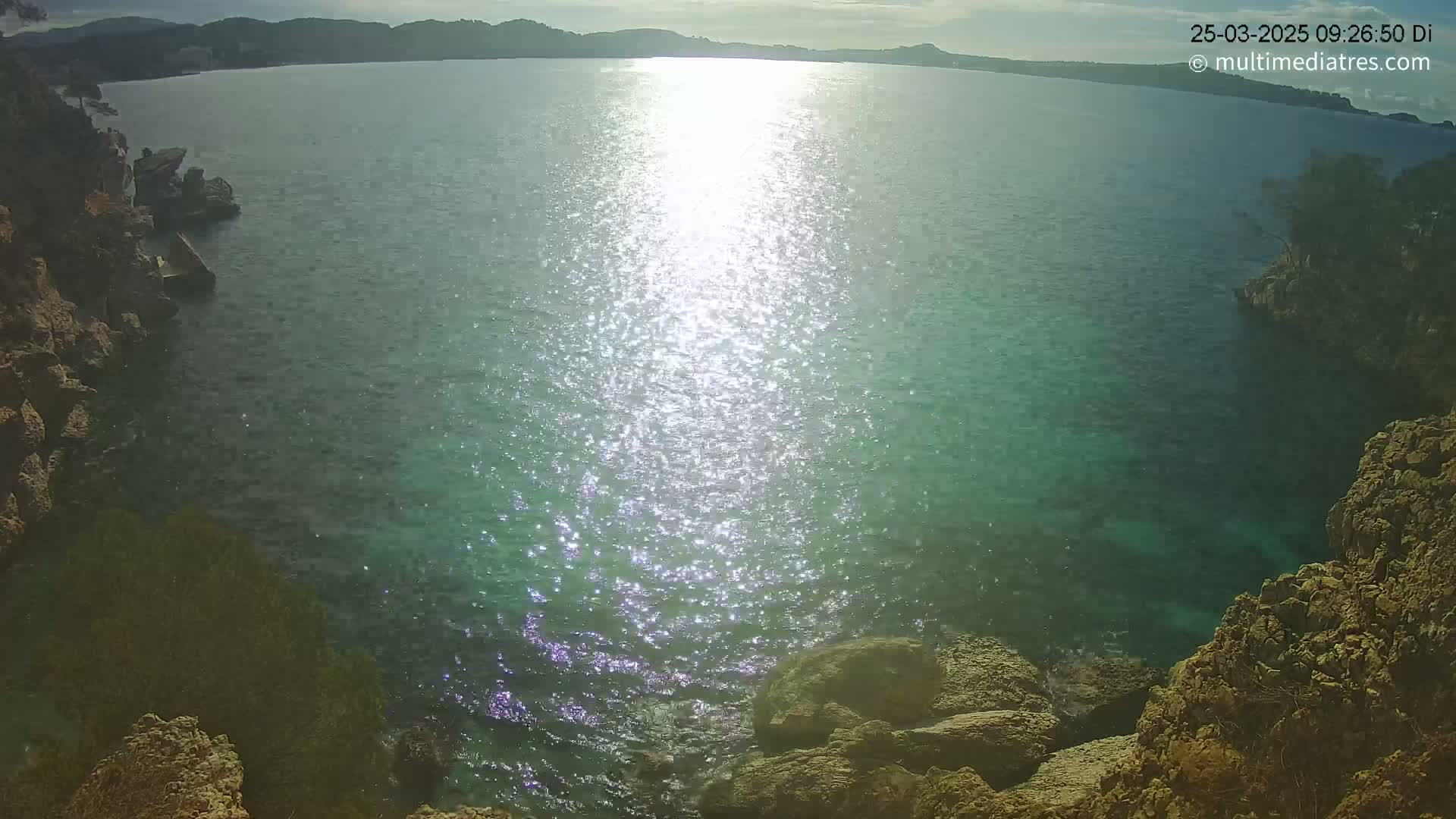 Rocky shores surround a calm, sparkling body of water on a partly sunny day.