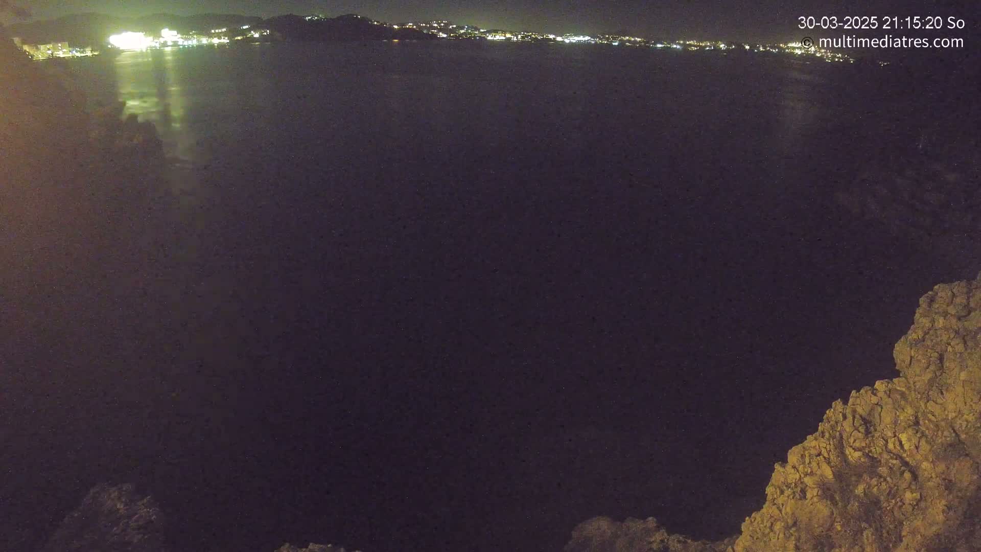 A nighttime view shows a dark body of water reflecting the lights of a distant city, with a rocky outcrop in the lower right corner.