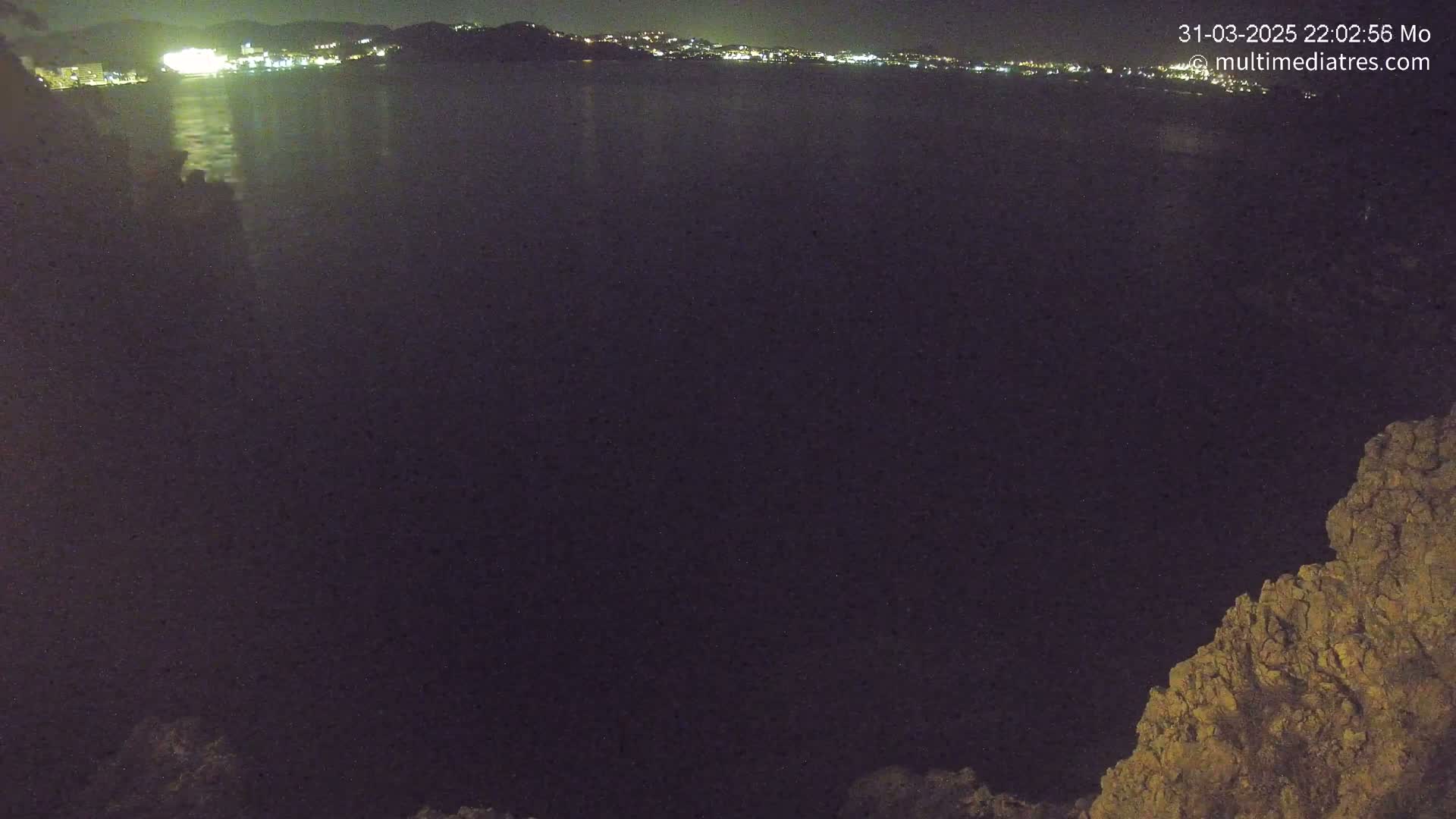 A nighttime view of a calm body of water reflecting the lights of a distant city, with a rocky outcrop in the lower right corner.