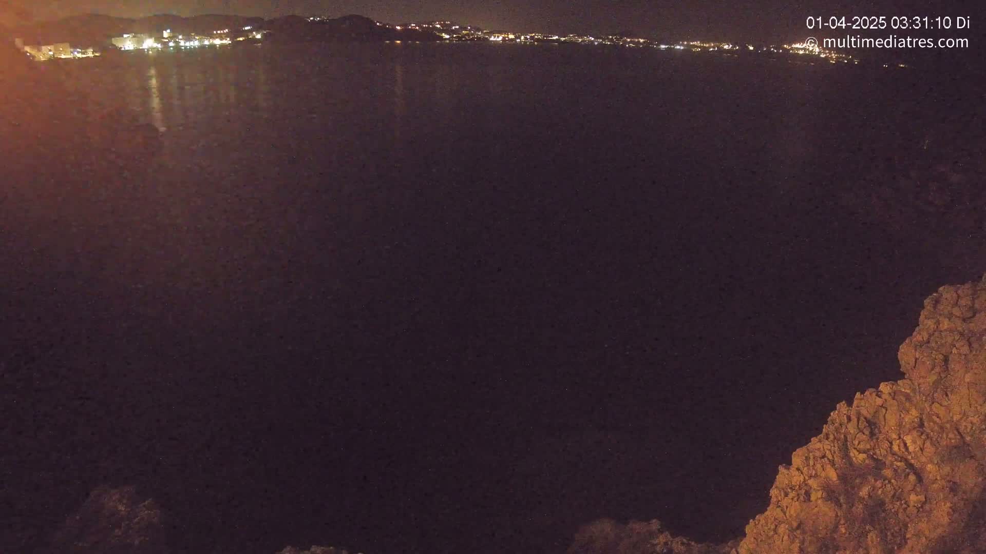 A nighttime, long shot of a dark body of water reflecting the lights of a distant town, with rocky terrain visible in the bottom right corner.