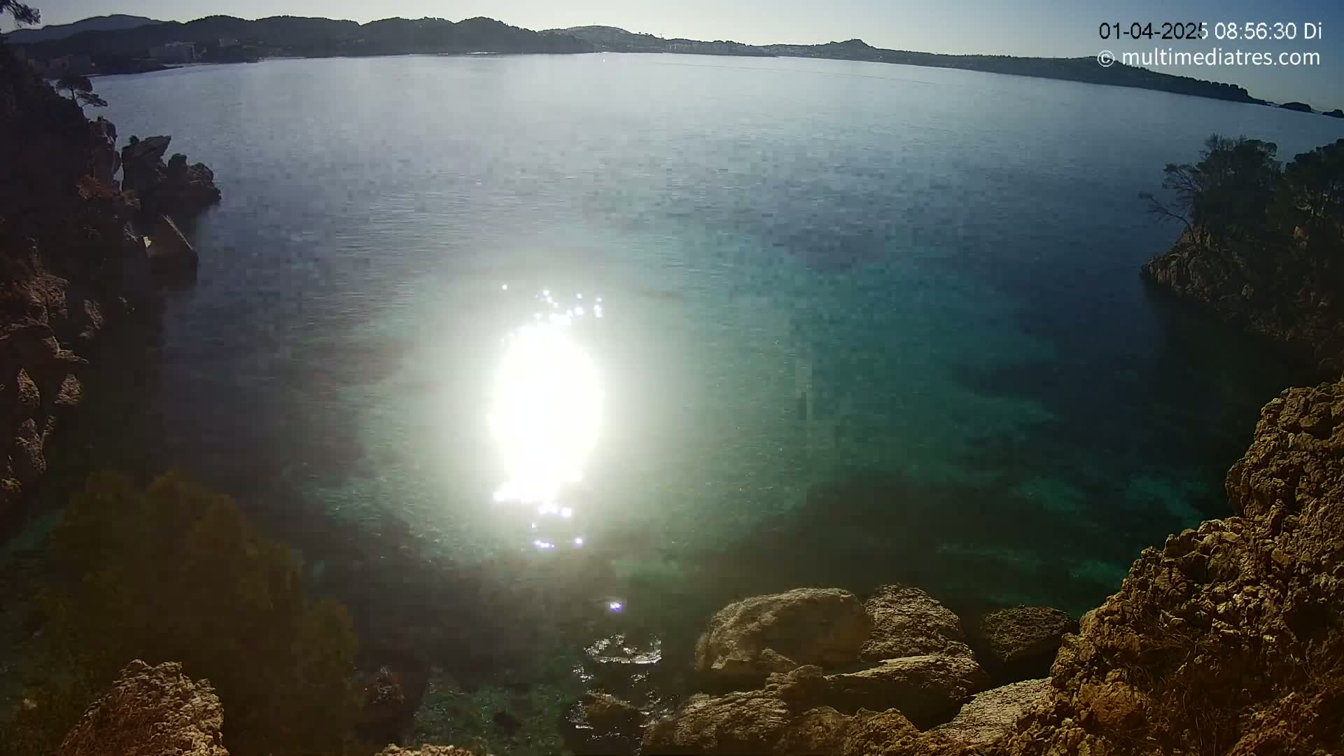 Sunlight glistens on the surface of clear, teal water nestled between rocky shorelines on a sunny day.