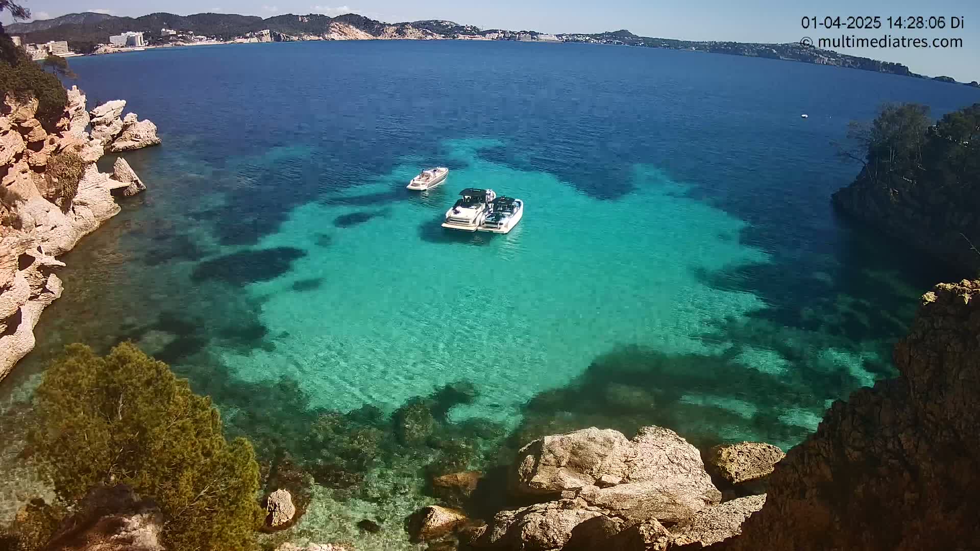 Two boats are anchored in a clear, turquoise cove surrounded by rocky cliffs and vegetation under a sunny sky.