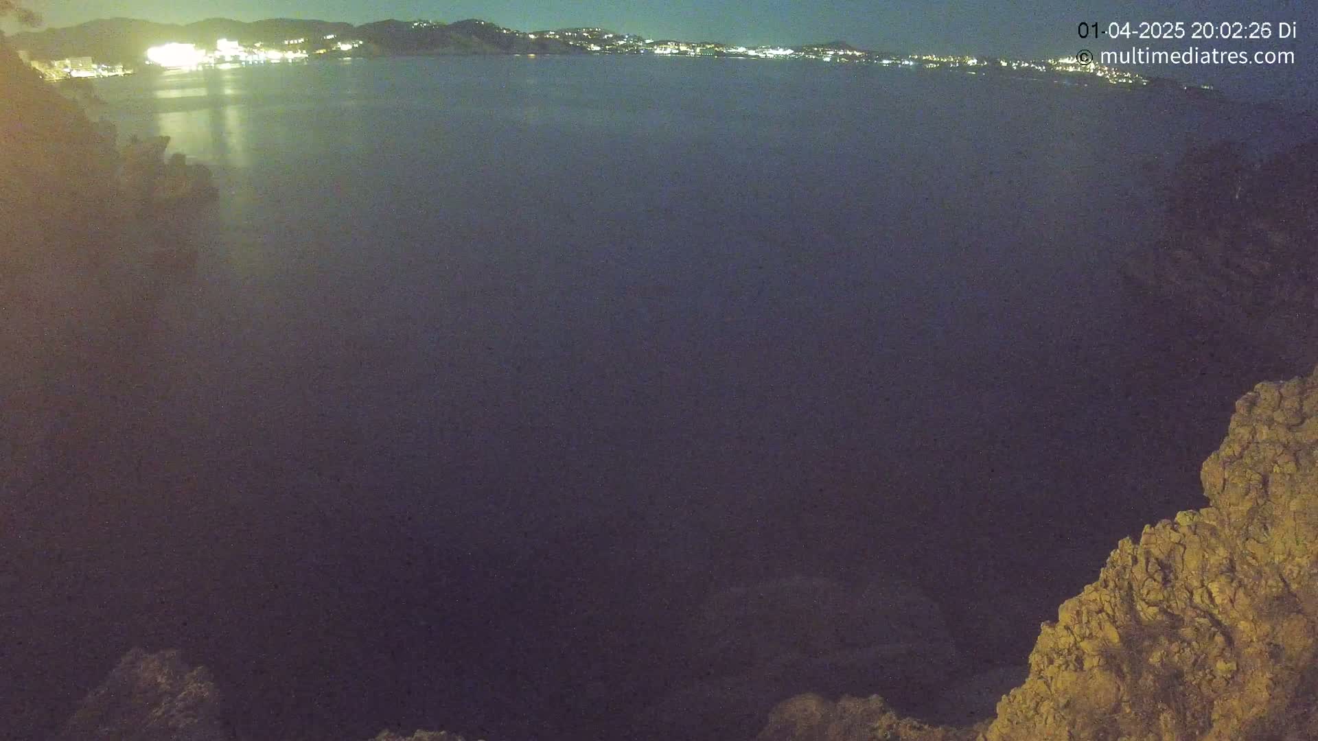 A nighttime view of a dark body of water with a distant, illuminated city skyline and rocky terrain in the foreground.