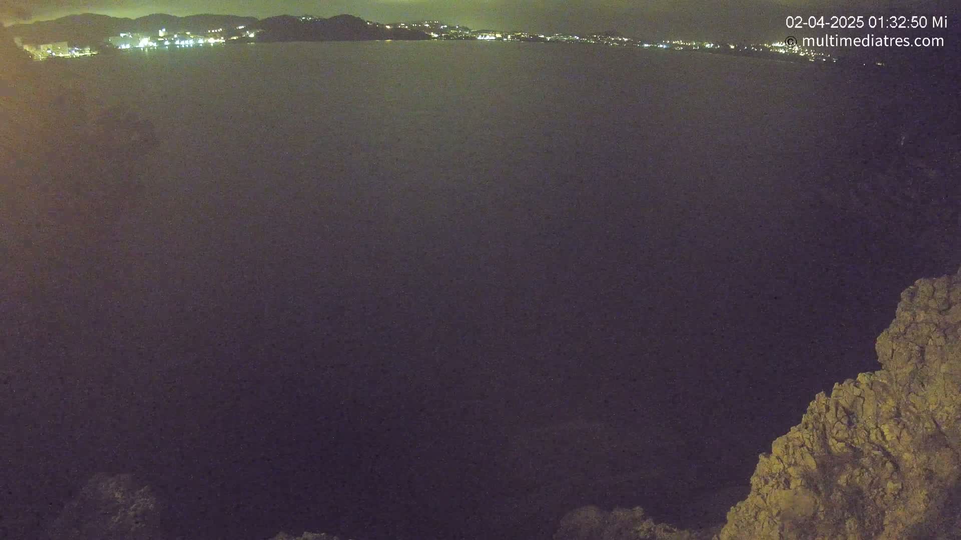 A dark body of water at night is seen from a high vantage point, with a distant city's lights visible along the shoreline and a rocky outcrop in the bottom right corner.