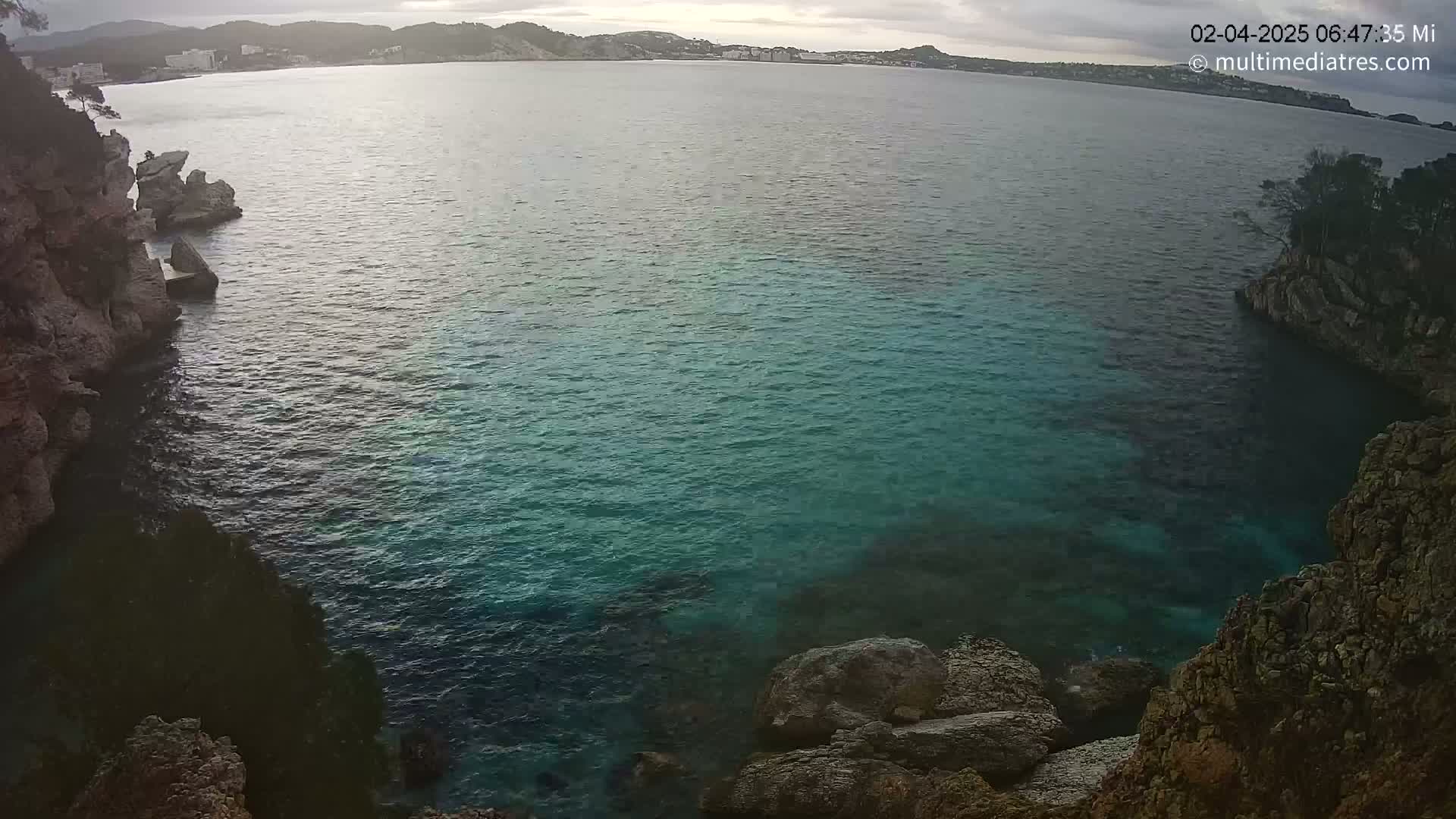 A tranquil cove with teal water is nestled between rocky cliffs under an overcast sky.