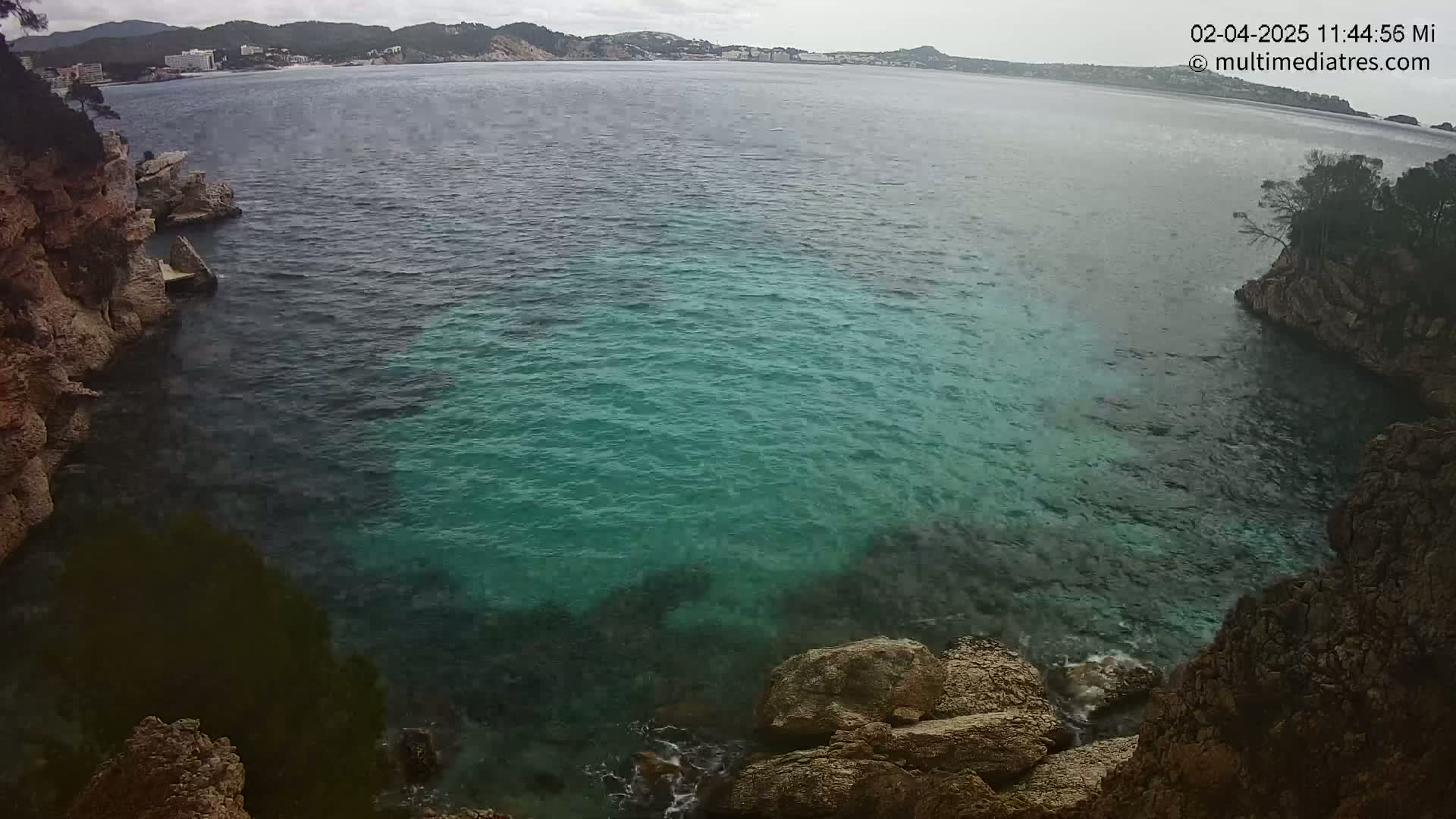 A cove of teal water is nestled between rocky cliffs, with a distant coastline visible under an overcast sky.