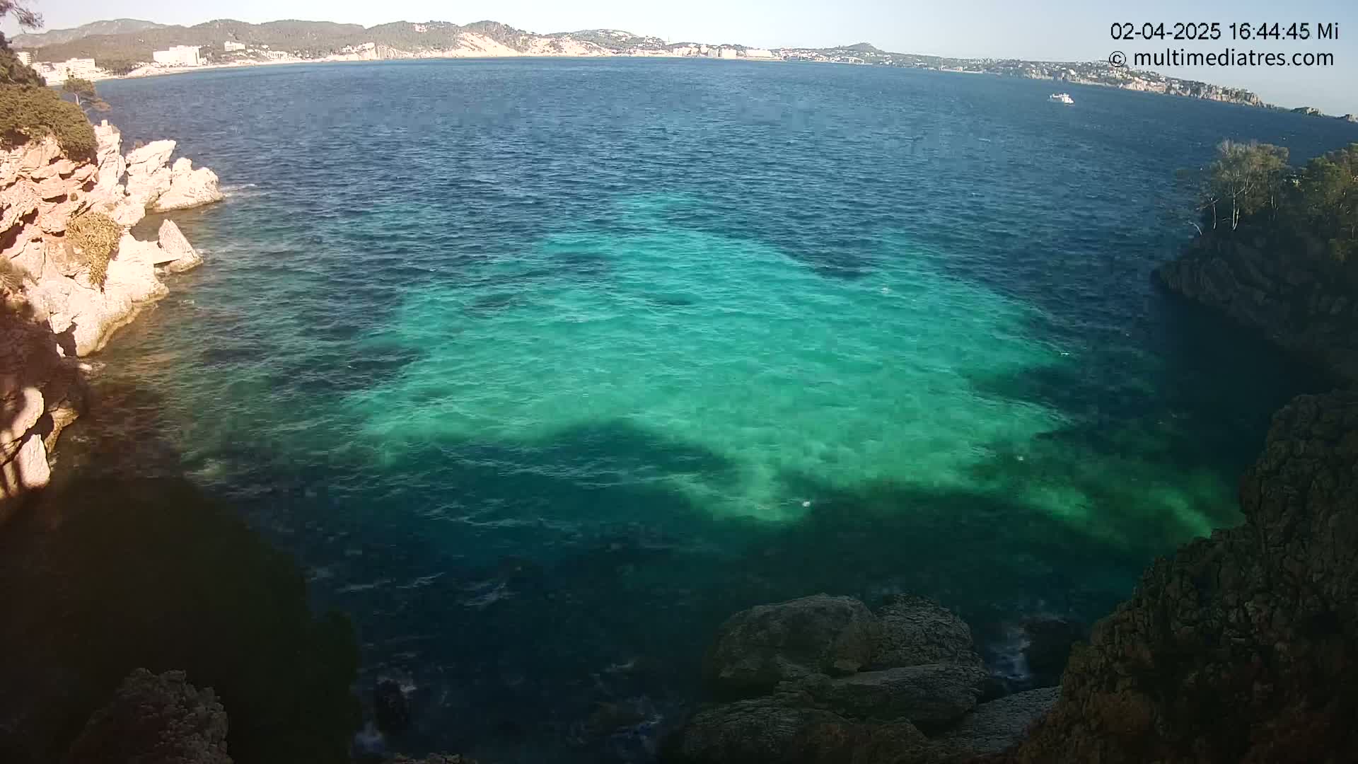 Clear, teal-colored water laps against rocky, tan-colored cliffs on a sunny day, with a distant coastline visible across the bay.